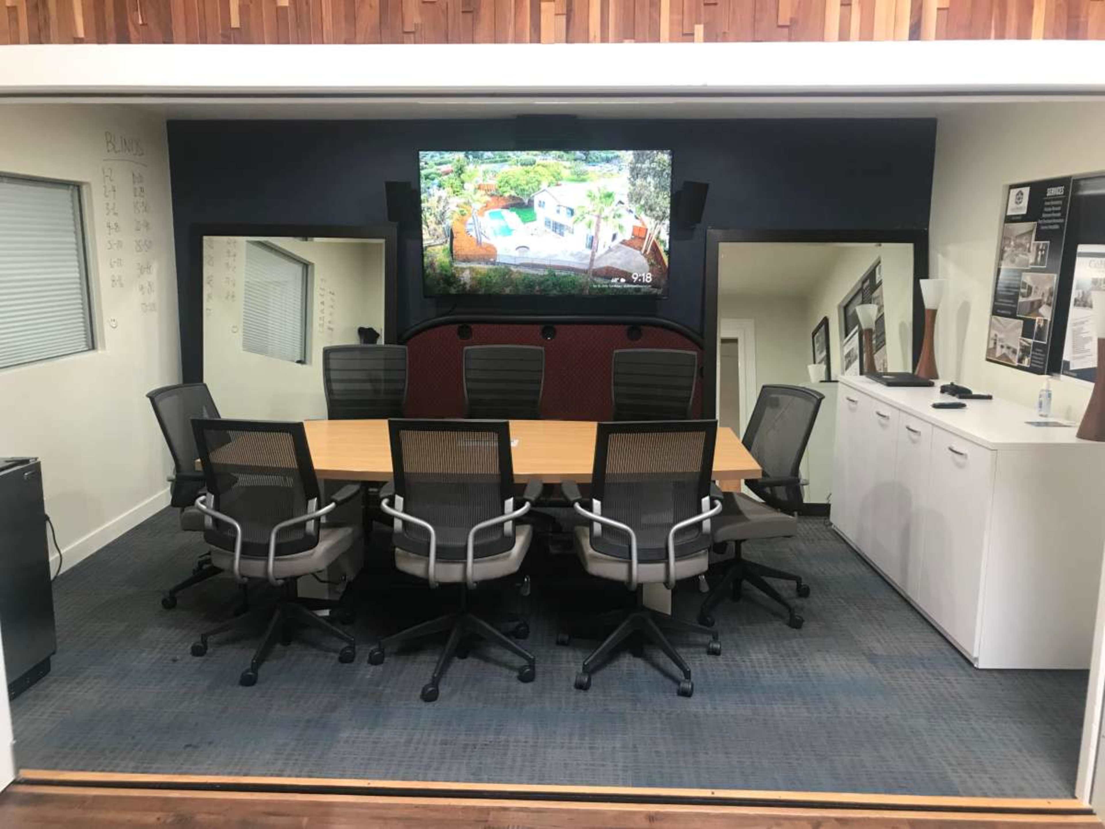 The image shows a modern conference room with a long wooden table surrounded by ergonomic chairs and a large television mounted on the wall.