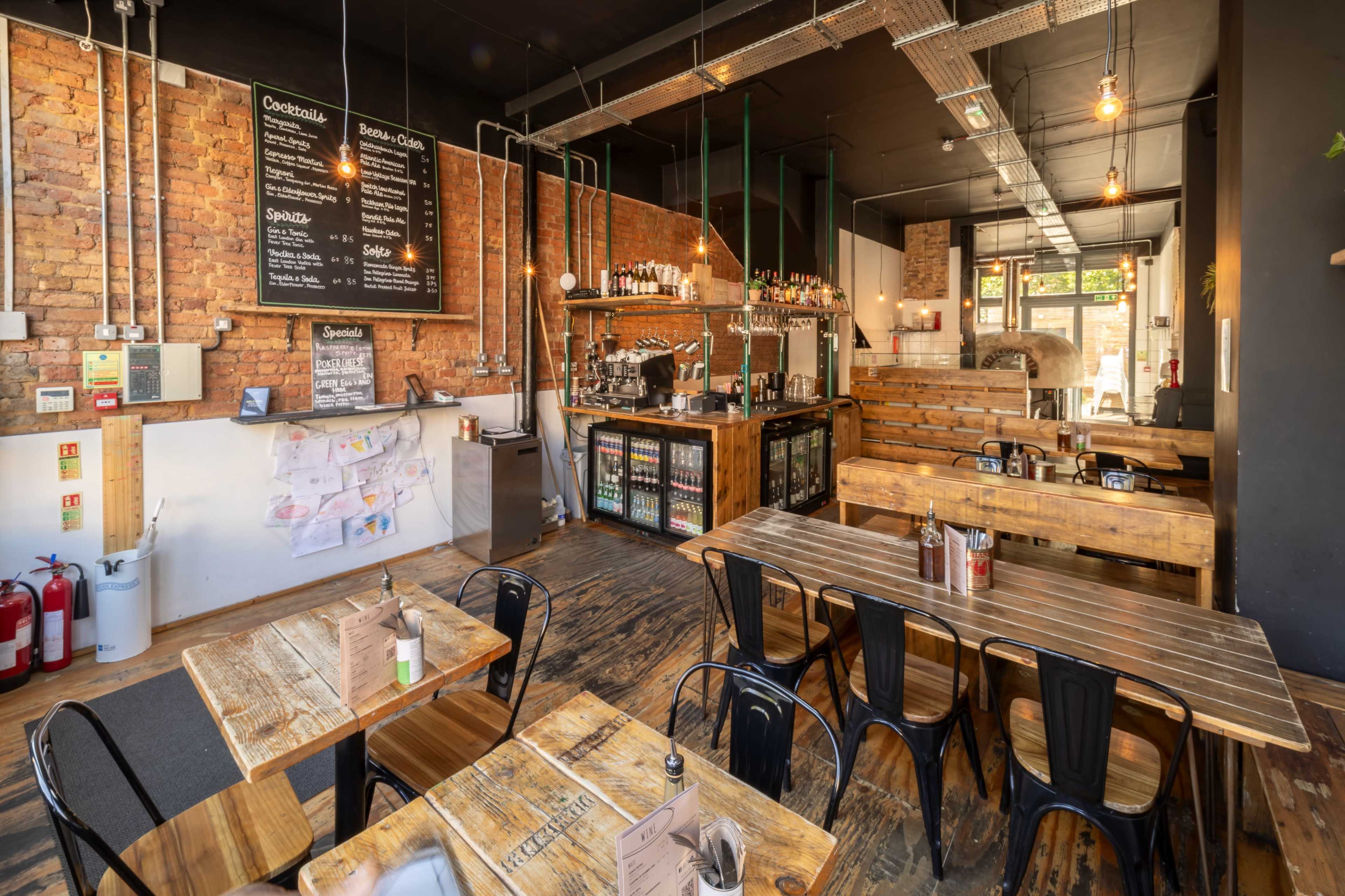 The image shows a modern restaurant interior featuring wooden tables and chairs, a bar area with shelves of drinks, and brick walls.