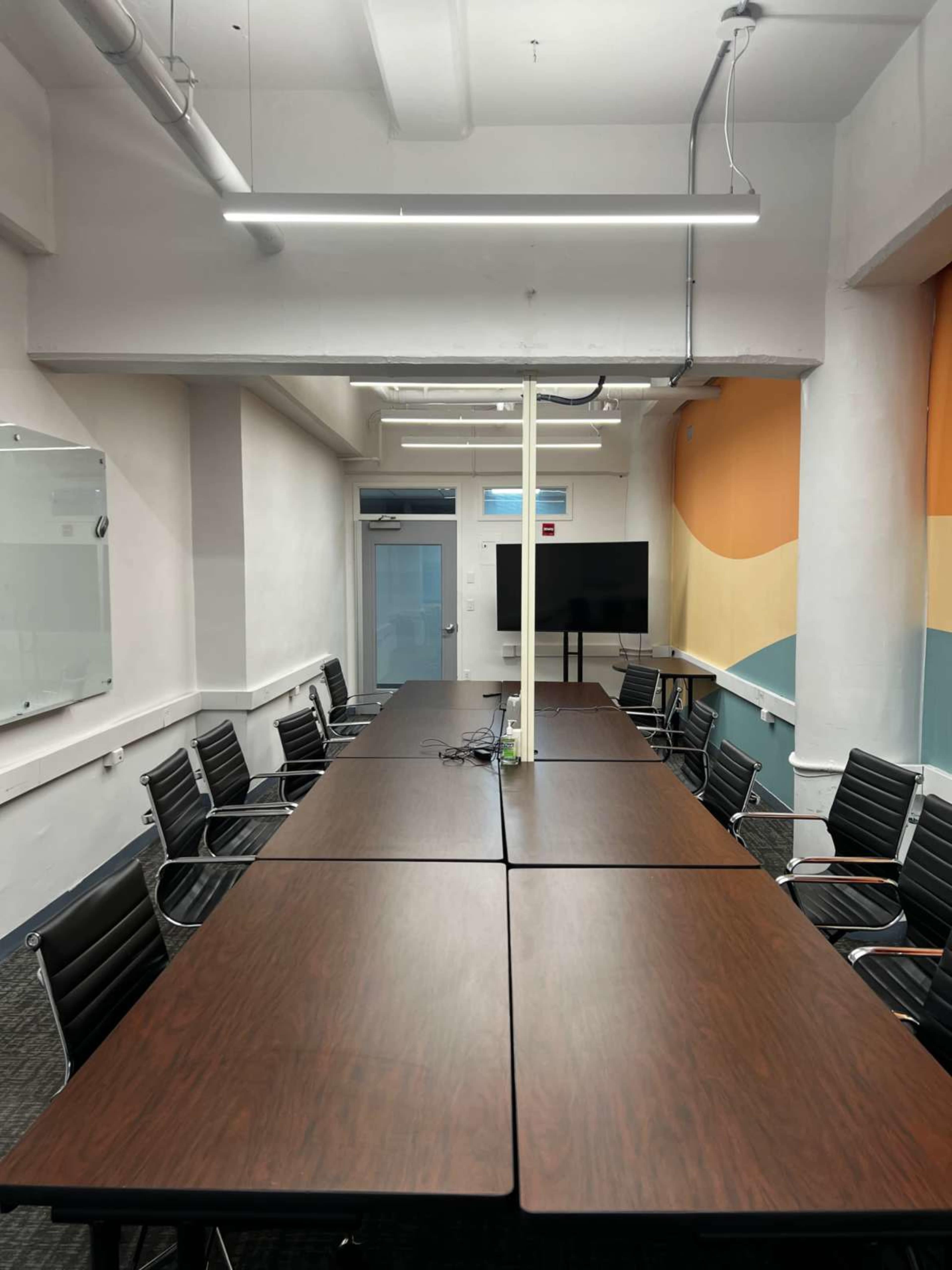 The image shows a modern conference room with a long wooden table, black chairs, and a large wall-mounted screen at one end.