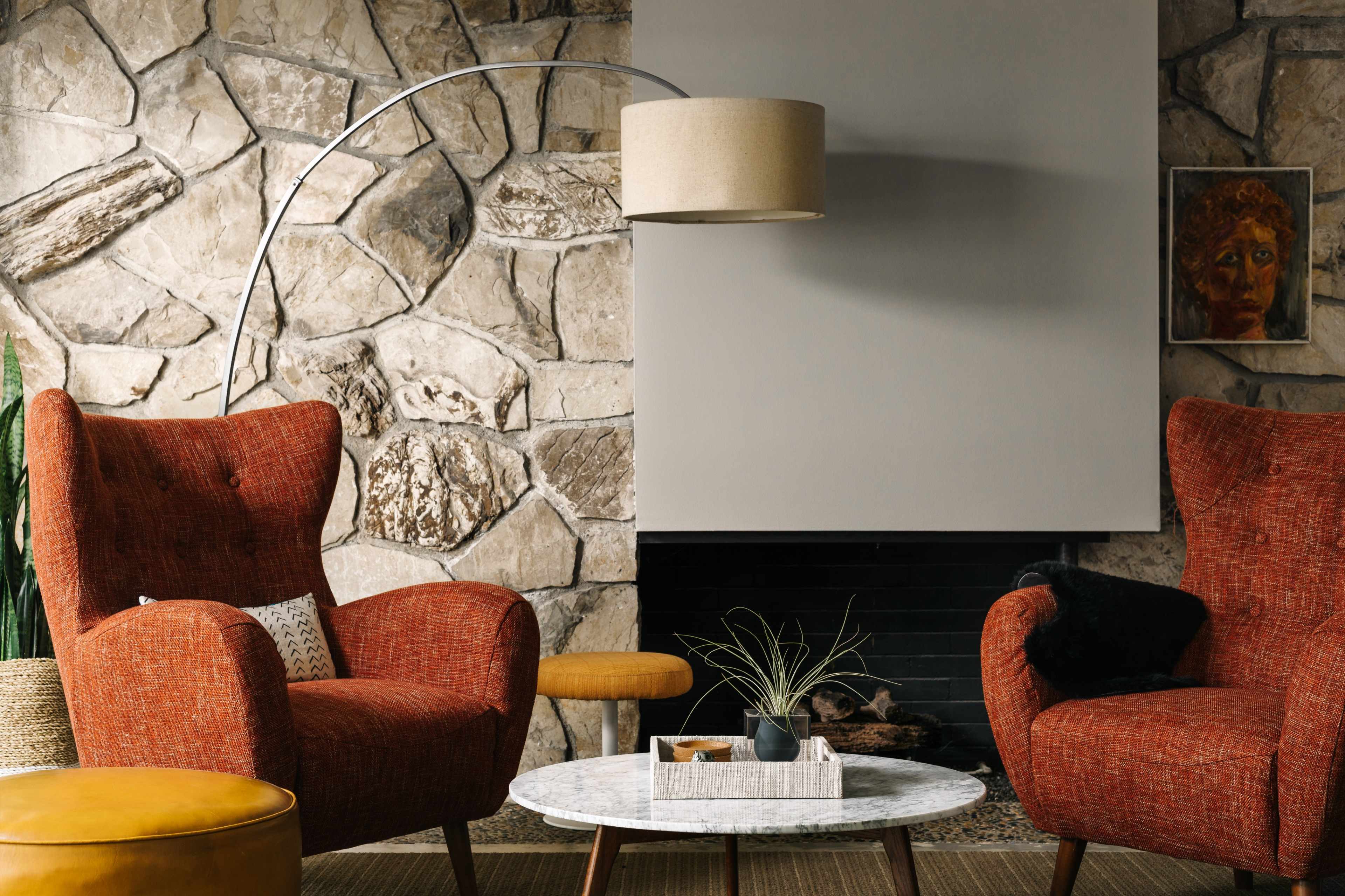 A cozy living room with two orange armchairs flanking a circular marble coffee table, set against a stone wall and featuring a floor lamp.