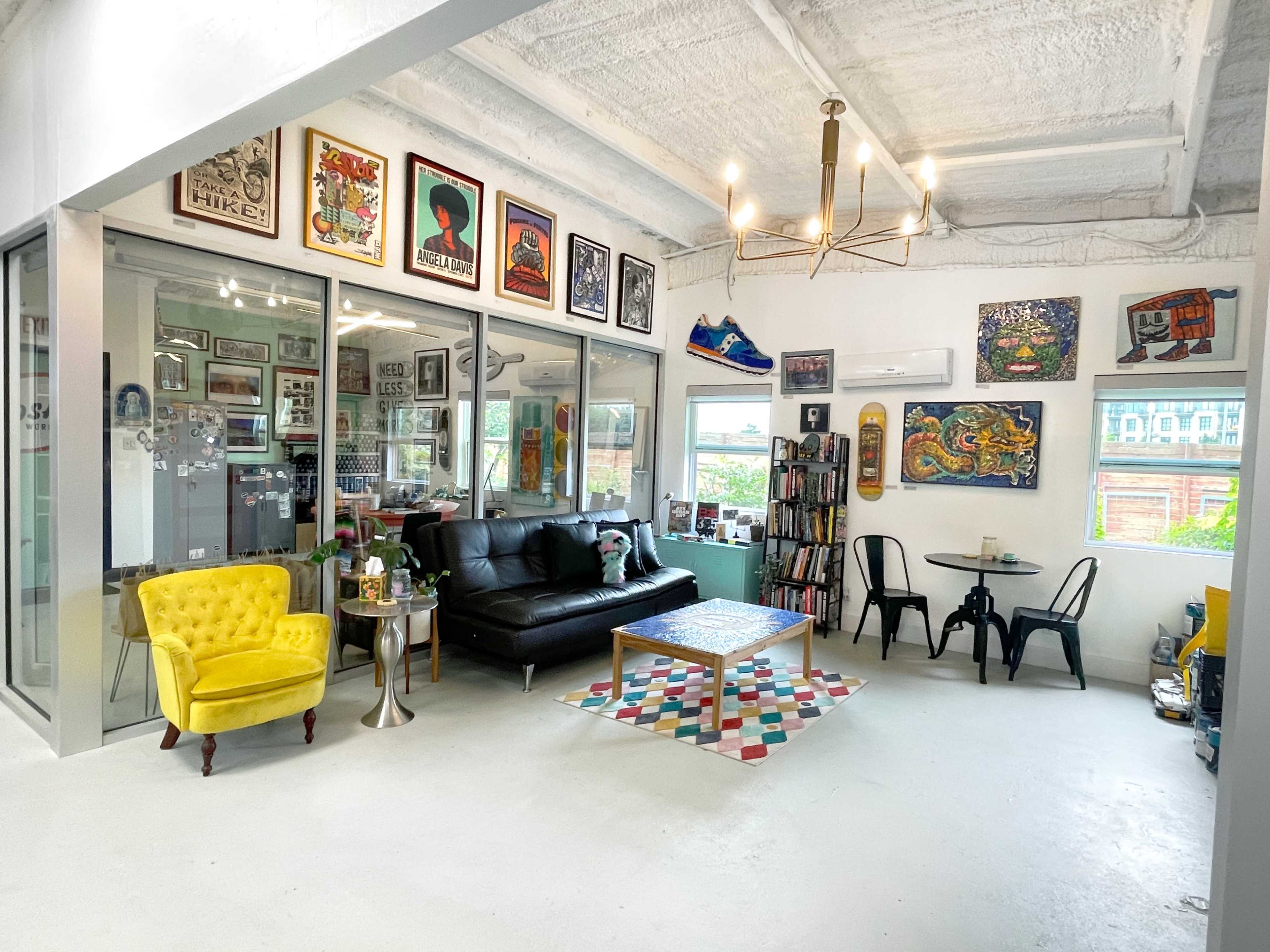 A bright, modern living room featuring a black sofa, a yellow armchair, a colorful rug, various wall artworks, and large windows letting in natural light.