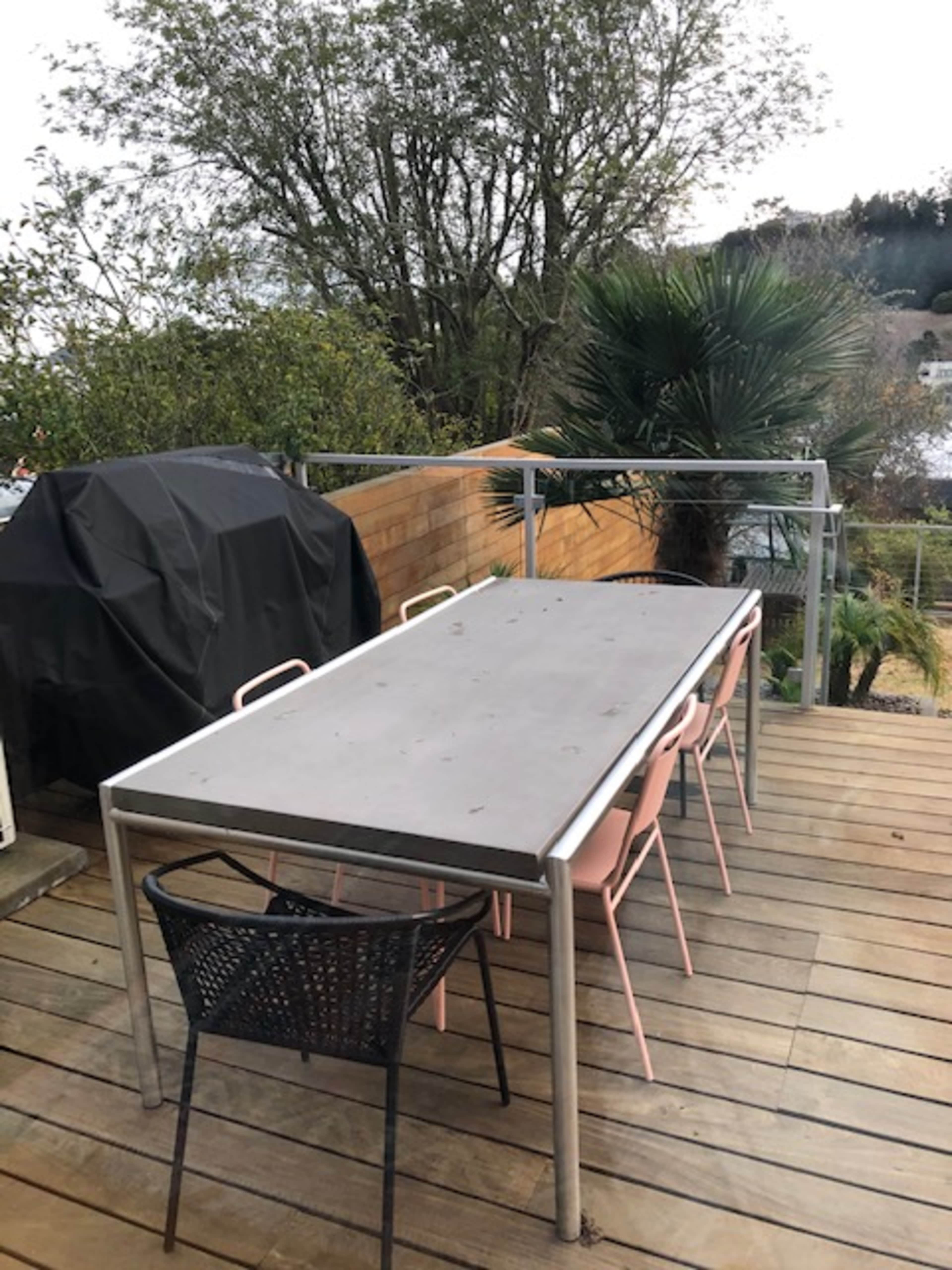 A wooden deck features a long metal table surrounded by chairs, with a grill covered in a black tarp nearby and trees in the background.