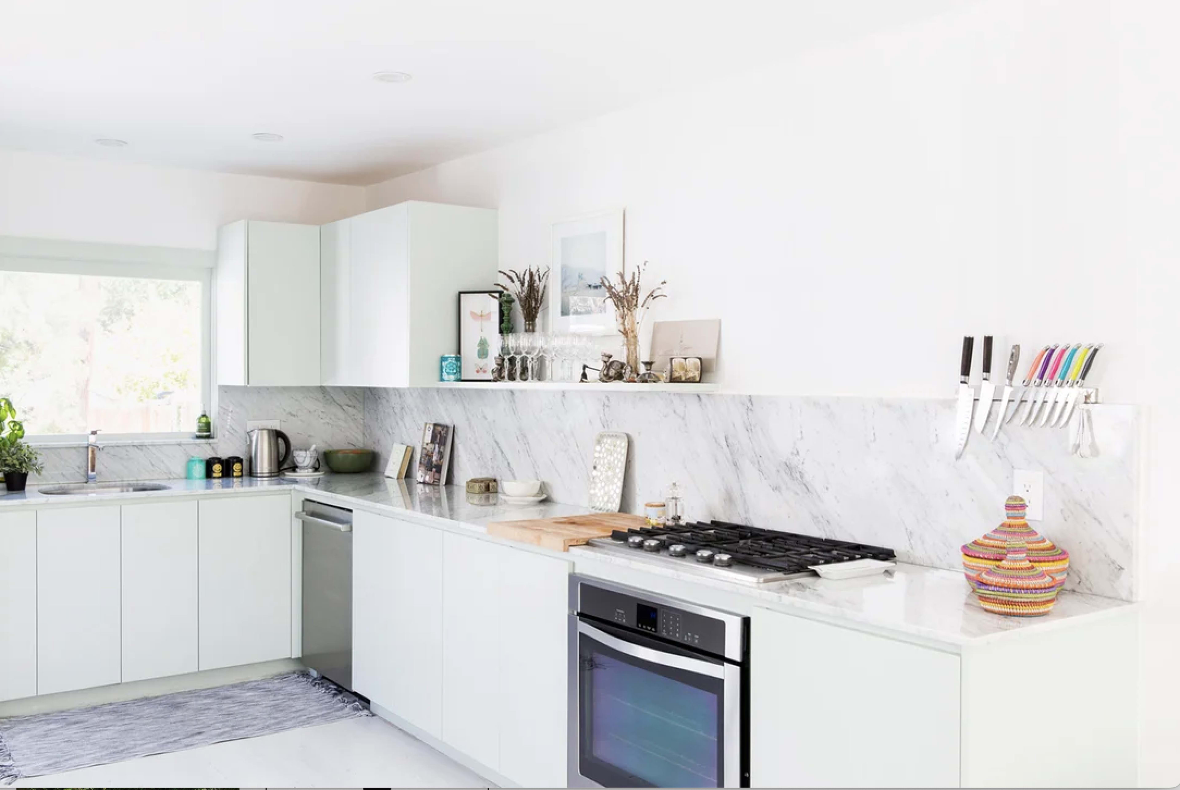 The image shows a modern kitchen with white cabinets, a marble backsplash, stainless steel appliances, and a wooden cutting board on the counter.
