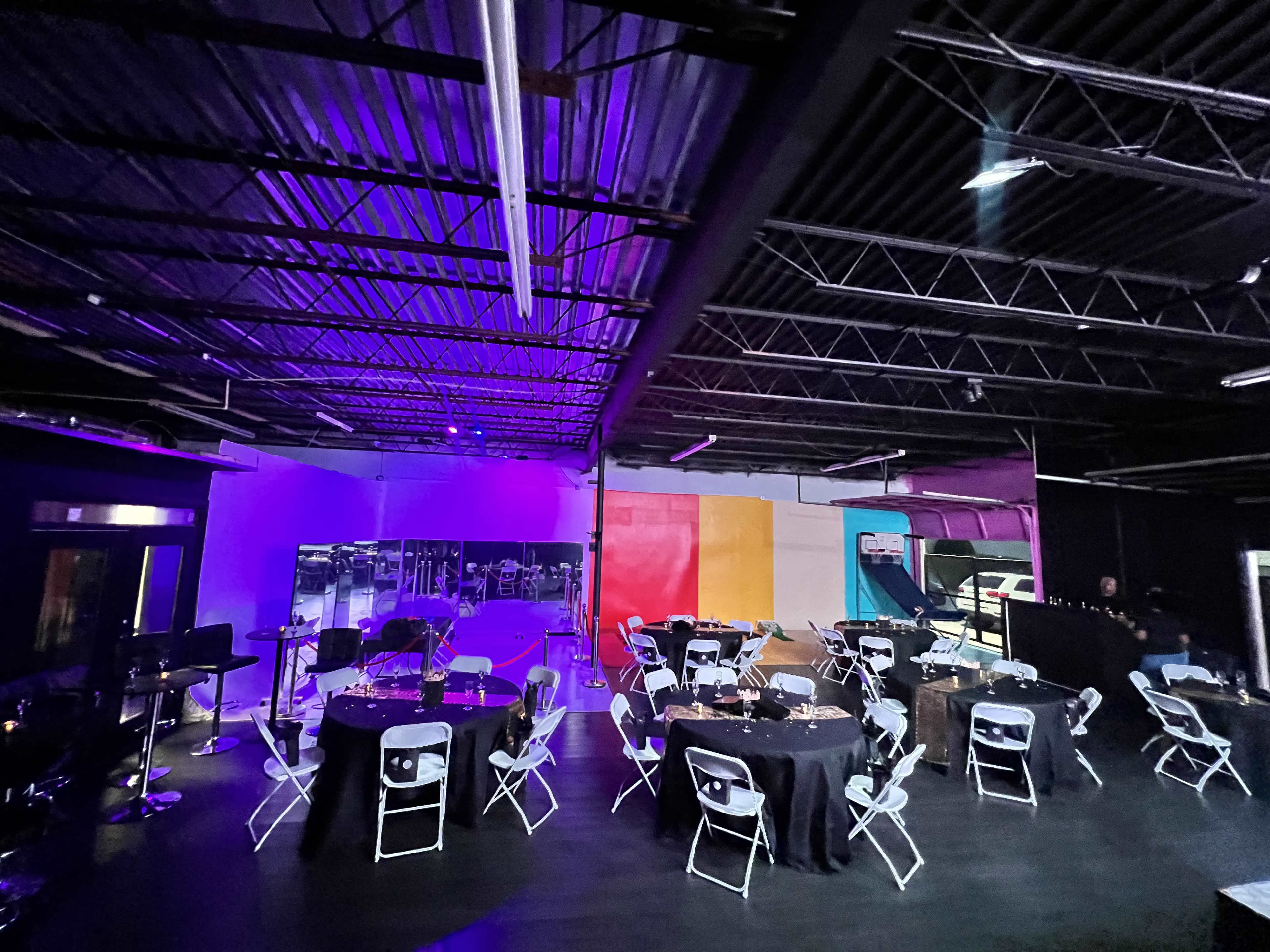 An event space with round tables and chairs set up under purple and blue lighting, featuring a dance floor area in the background.