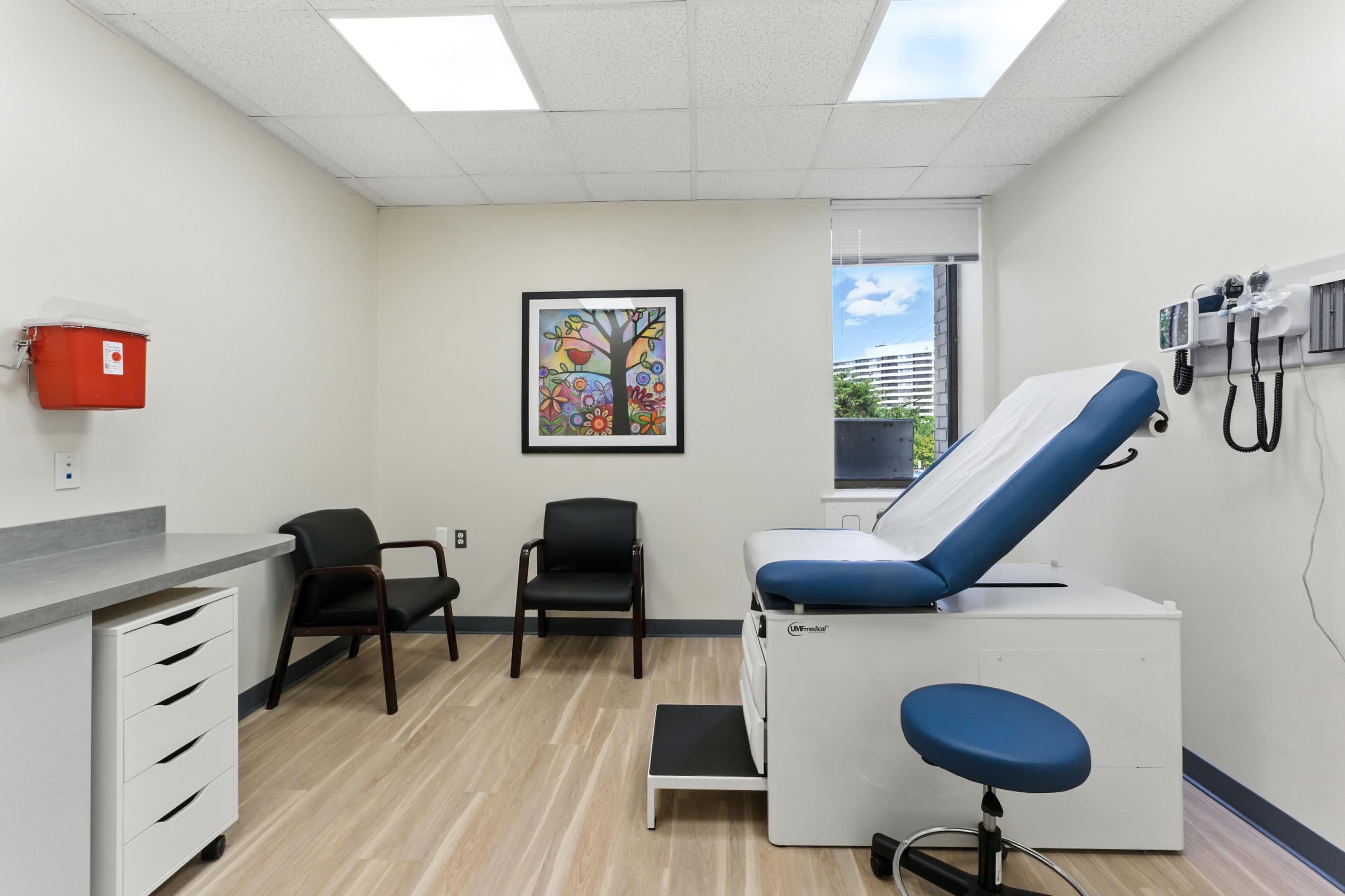 The image shows a medical examination room with a patient examination table, two chairs, a colorful wall art piece, and a window overlooking a building.