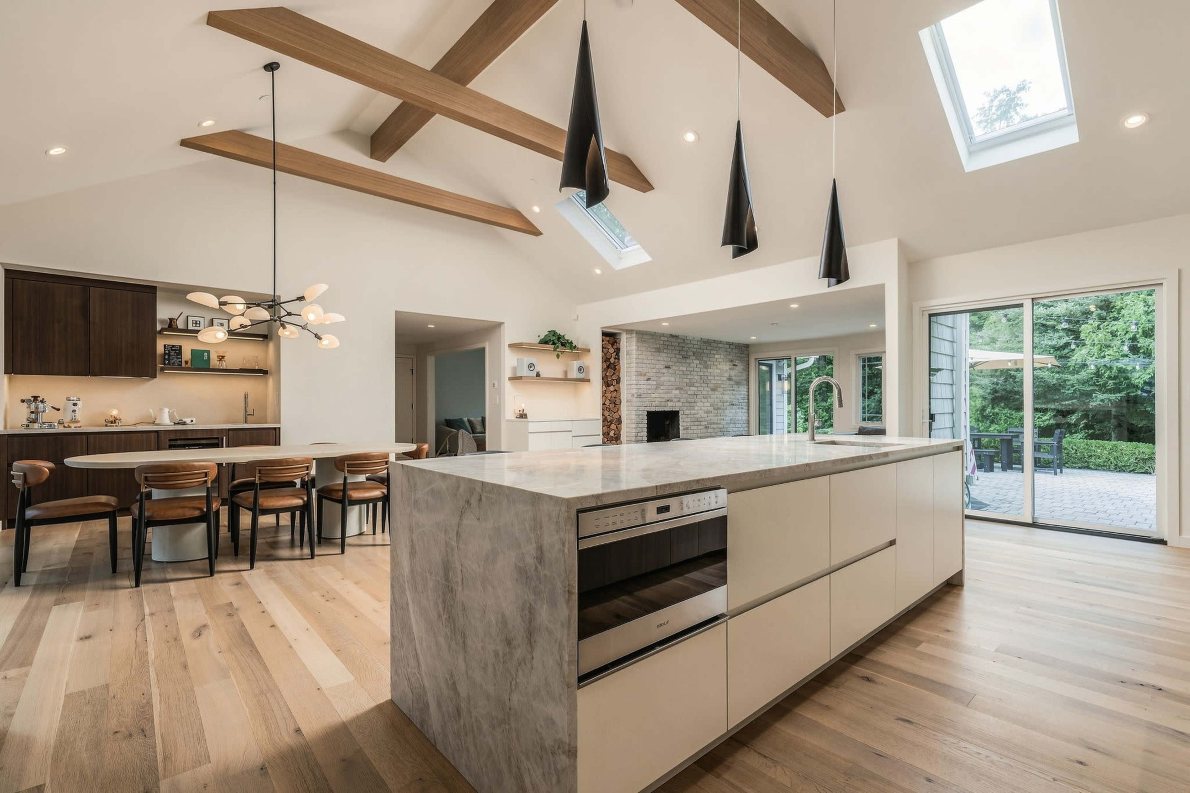 The image shows a modern kitchen with a large island countertop, wooden beams, and an adjacent dining area featuring a table and chairs.