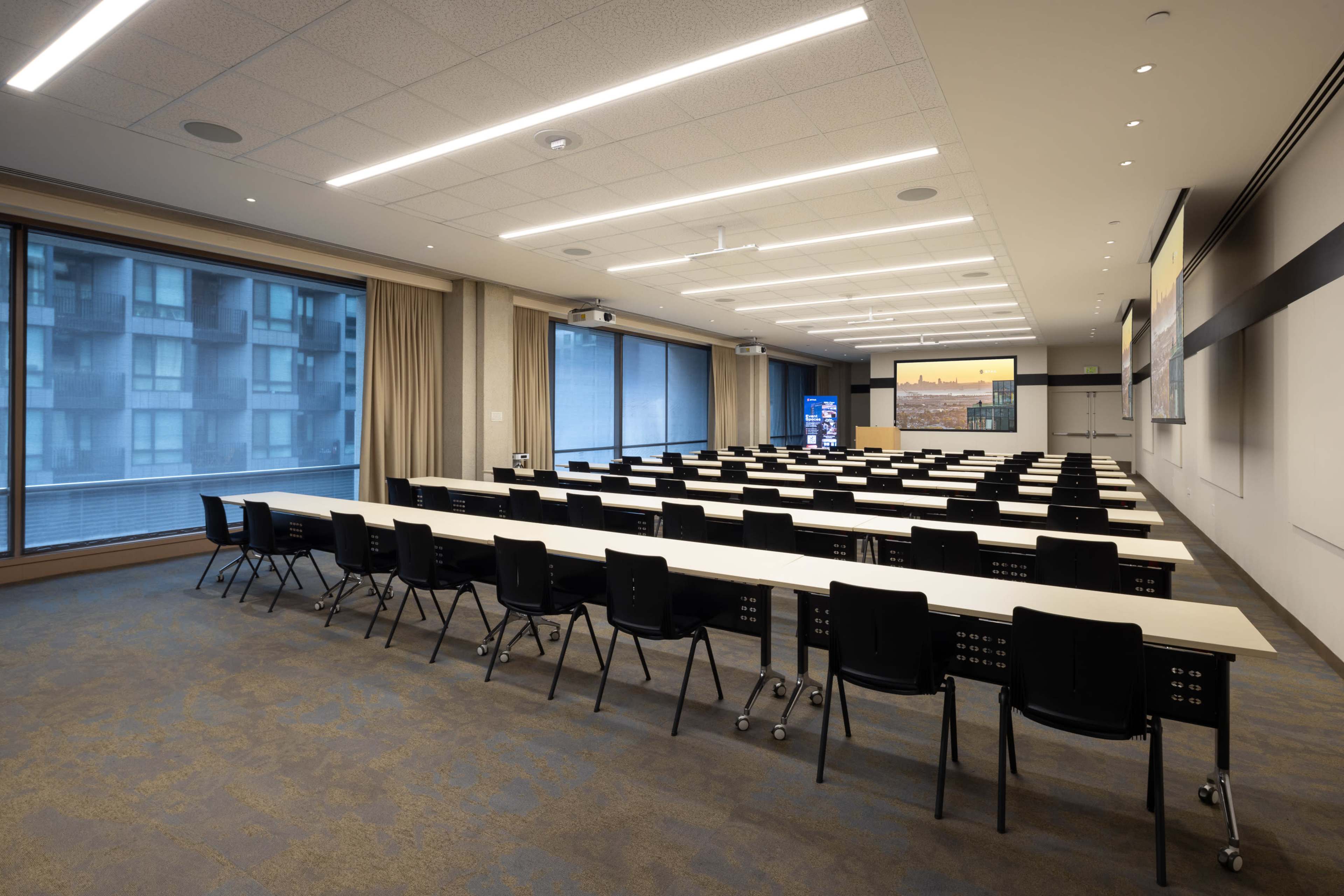A modern conference room features rows of tables and chairs, large windows, and a screen for presentations.