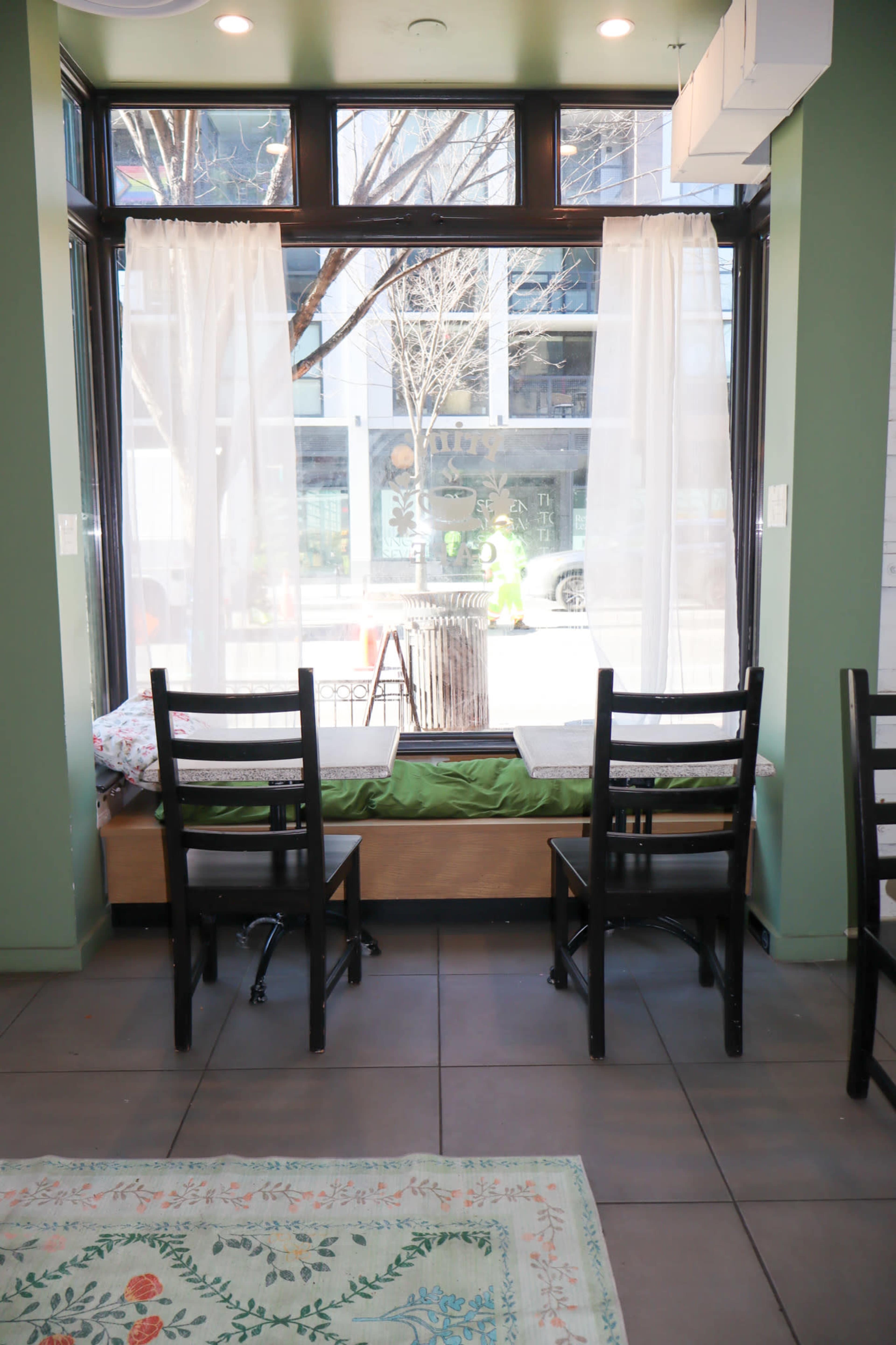 The image shows a bright indoor seating area with two empty chairs facing a window, featuring white curtains and a view of a street outside.