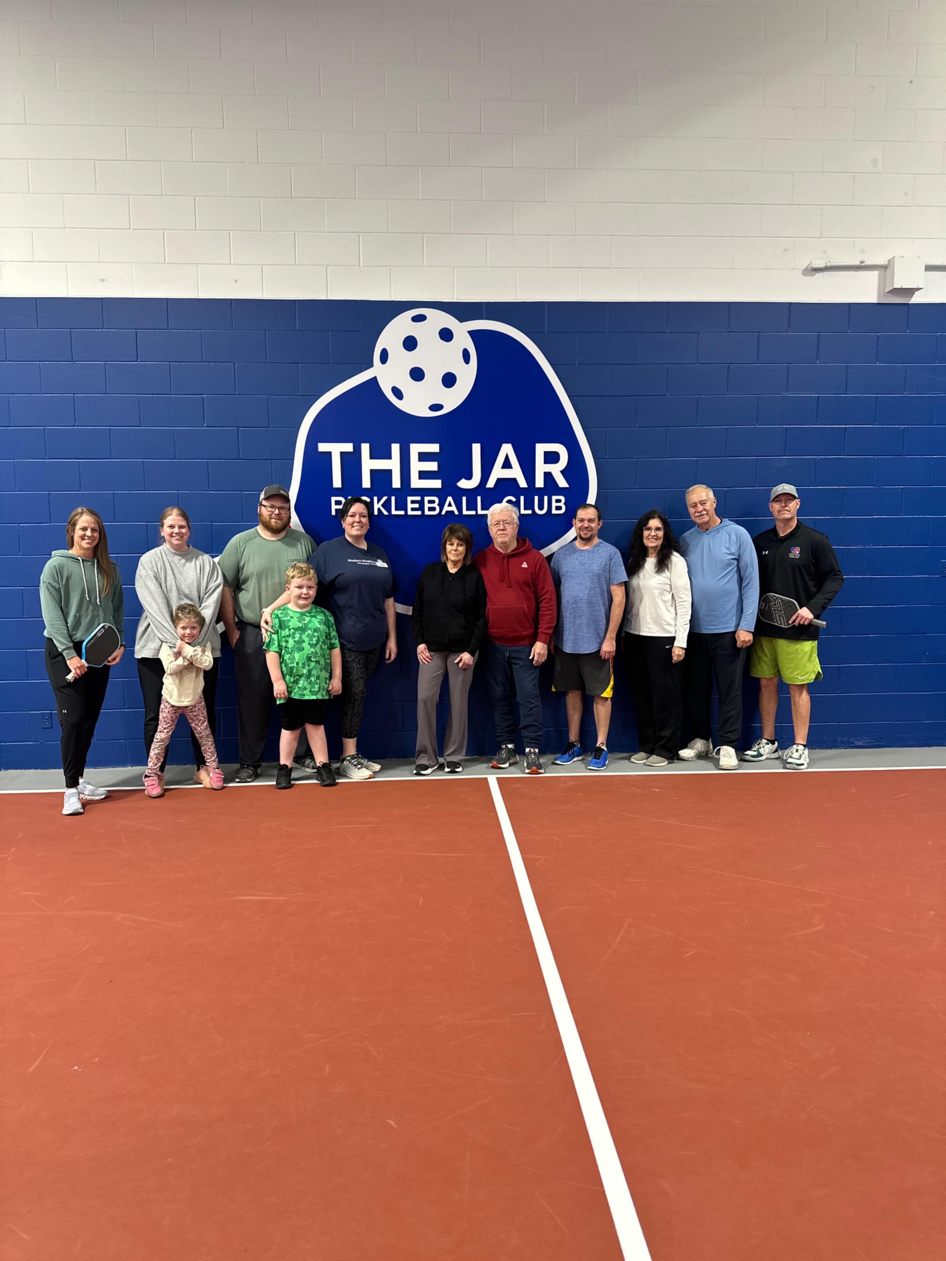 A group of people poses for a photo in front of a wall featuring the logo of "The Jar Pickleball Club" inside a sports facility.