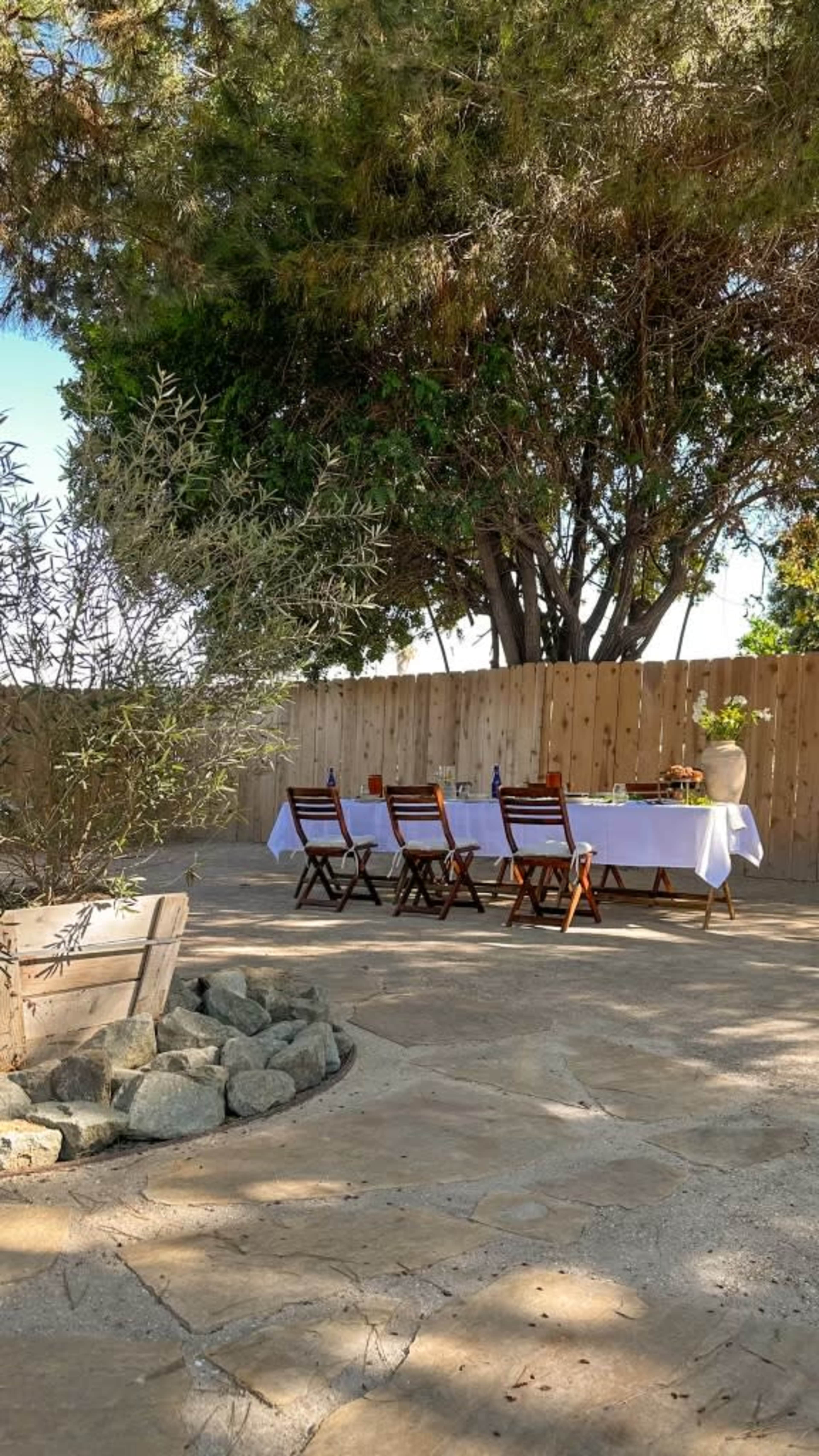 A long table is set with a white tablecloth and surrounded by wooden chairs in a shaded outdoor area with trees and a wooden fence.