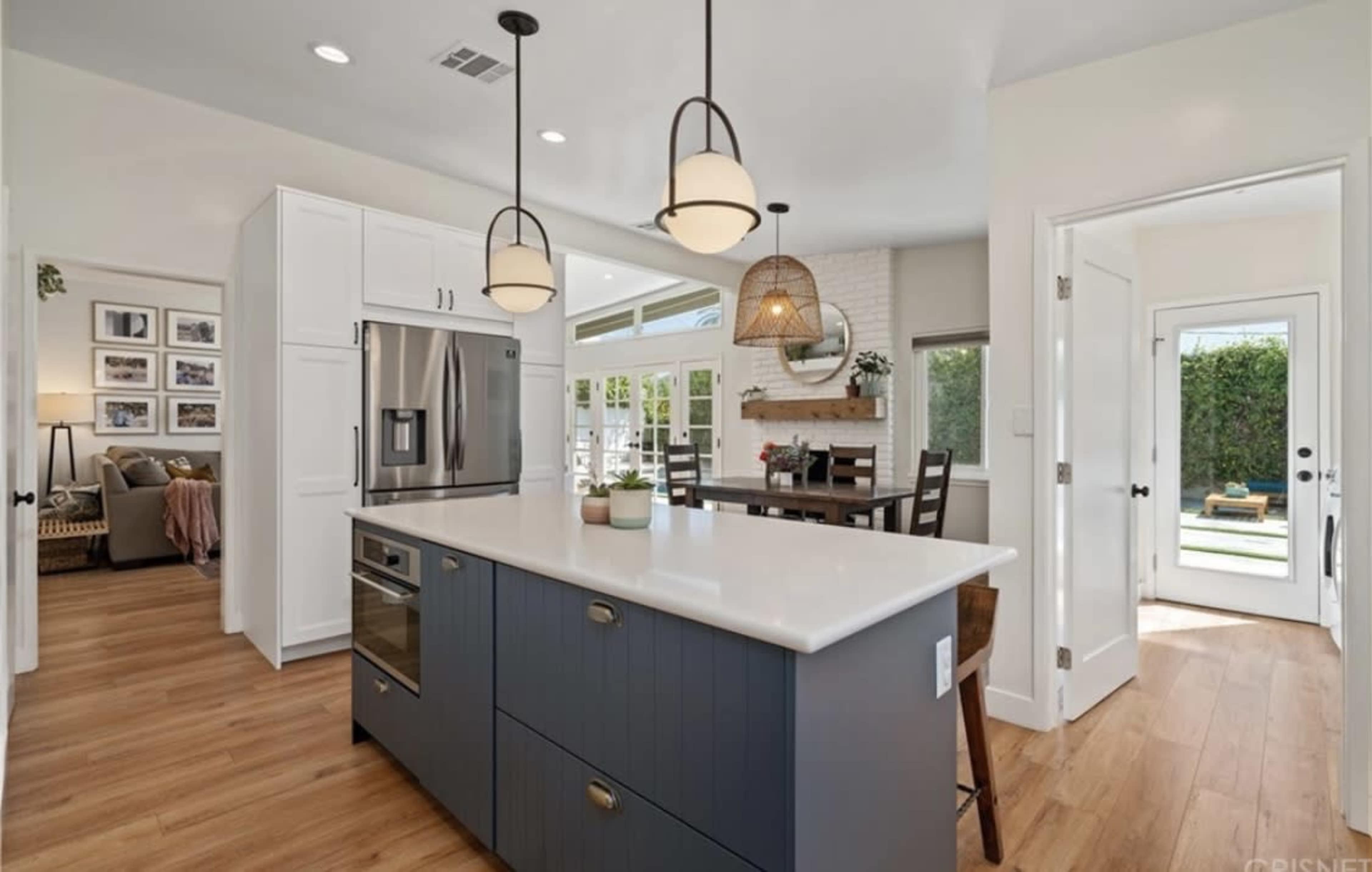 The image shows a modern kitchen with a central island featuring a built-in oven and a white countertop, connected to a dining area and a door leading to an outdoor space.