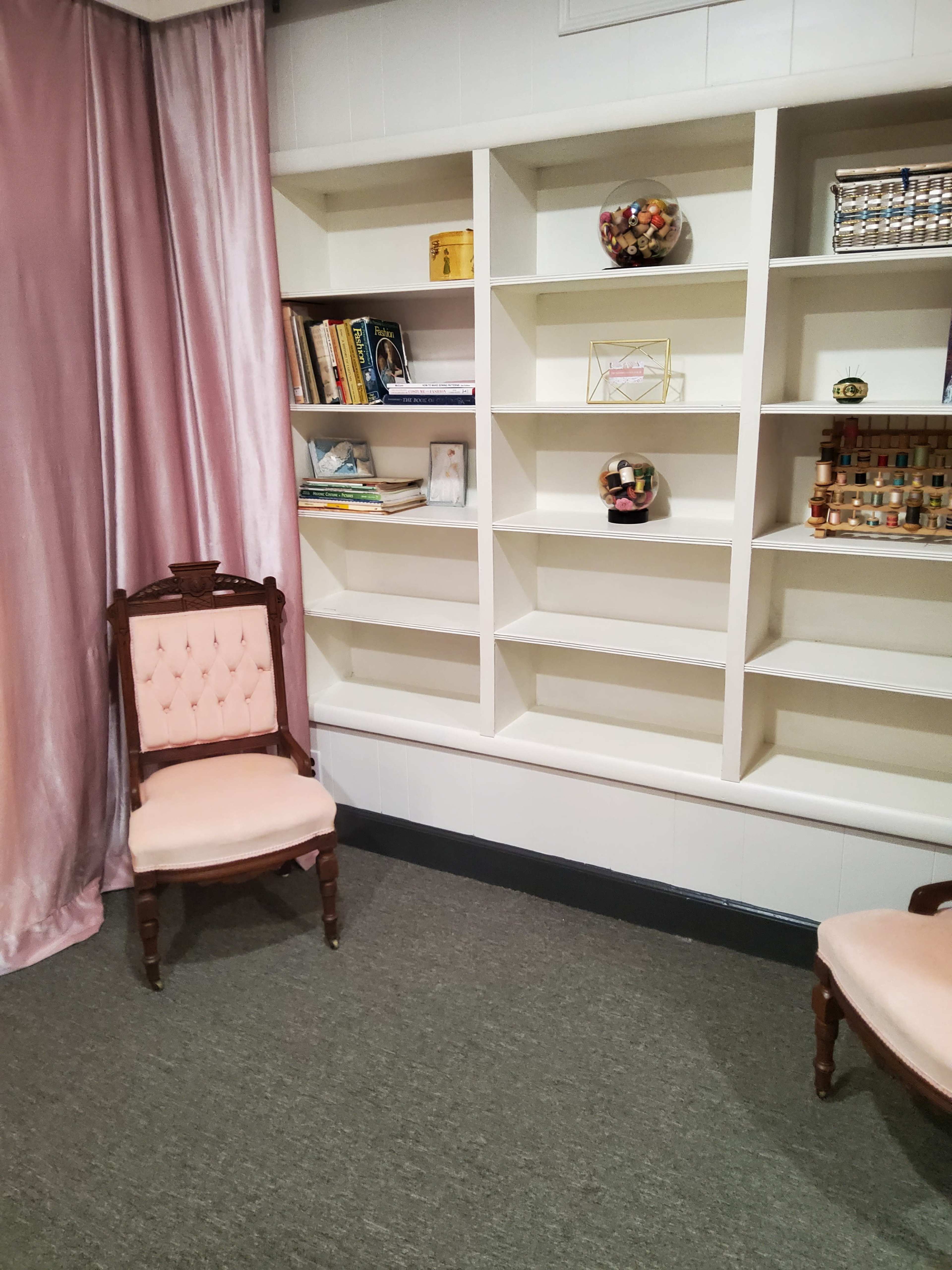 A room with two upholstered chairs and a white shelving unit displaying various objects and books, alongside a pink curtain.