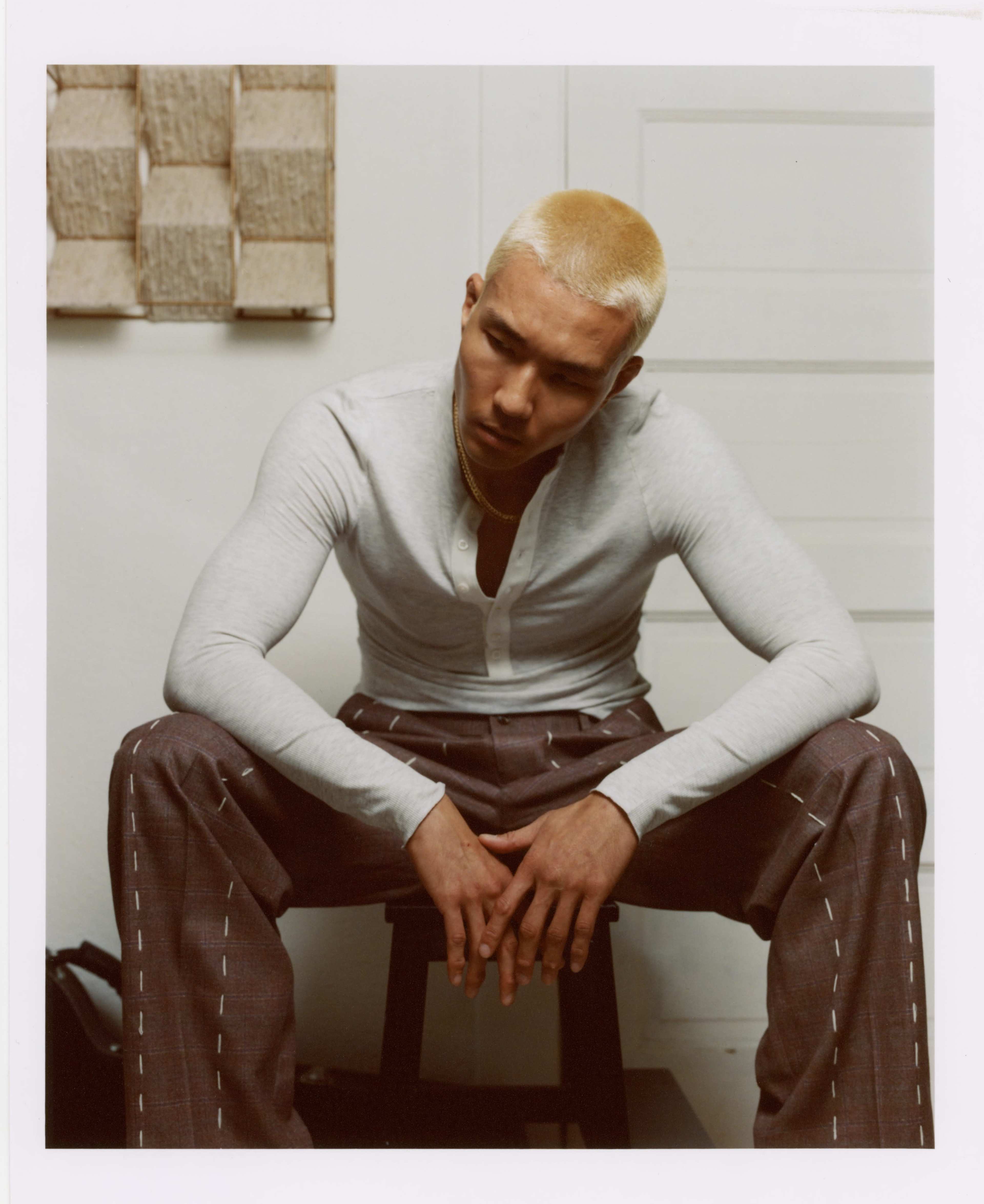 A young man with bleached hair sits on a stool, wearing a light gray long-sleeve shirt and patterned pants, with his hands clasped together in front of him.