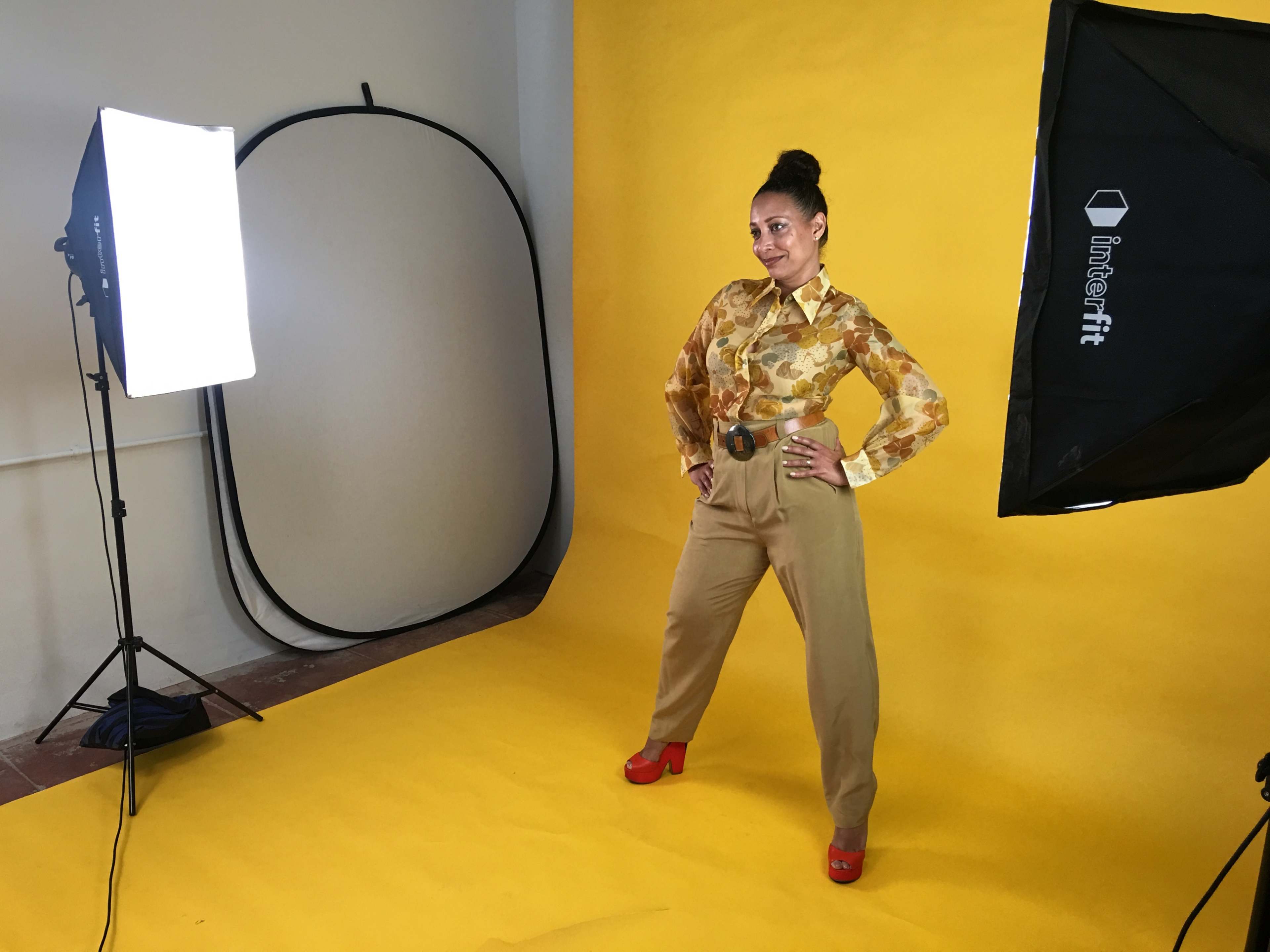 A model poses confidently in a brightly lit studio with a yellow backdrop, wearing a patterned blouse and tan pants.