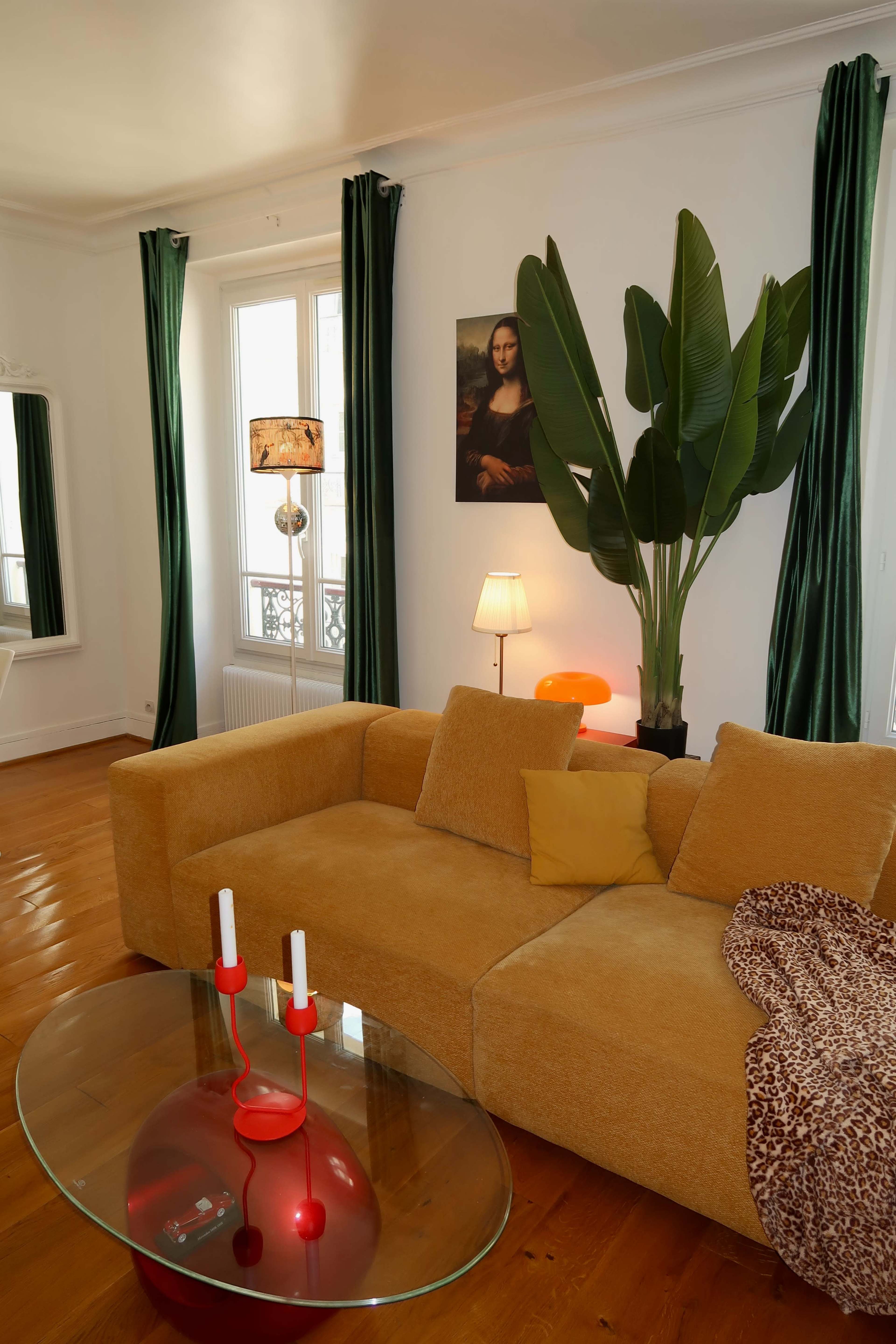 A cozy living room features a yellow curved sofa, a glass coffee table with red accents, large green plants, and tall curtains with a framed painting of the Mona Lisa on the wall.