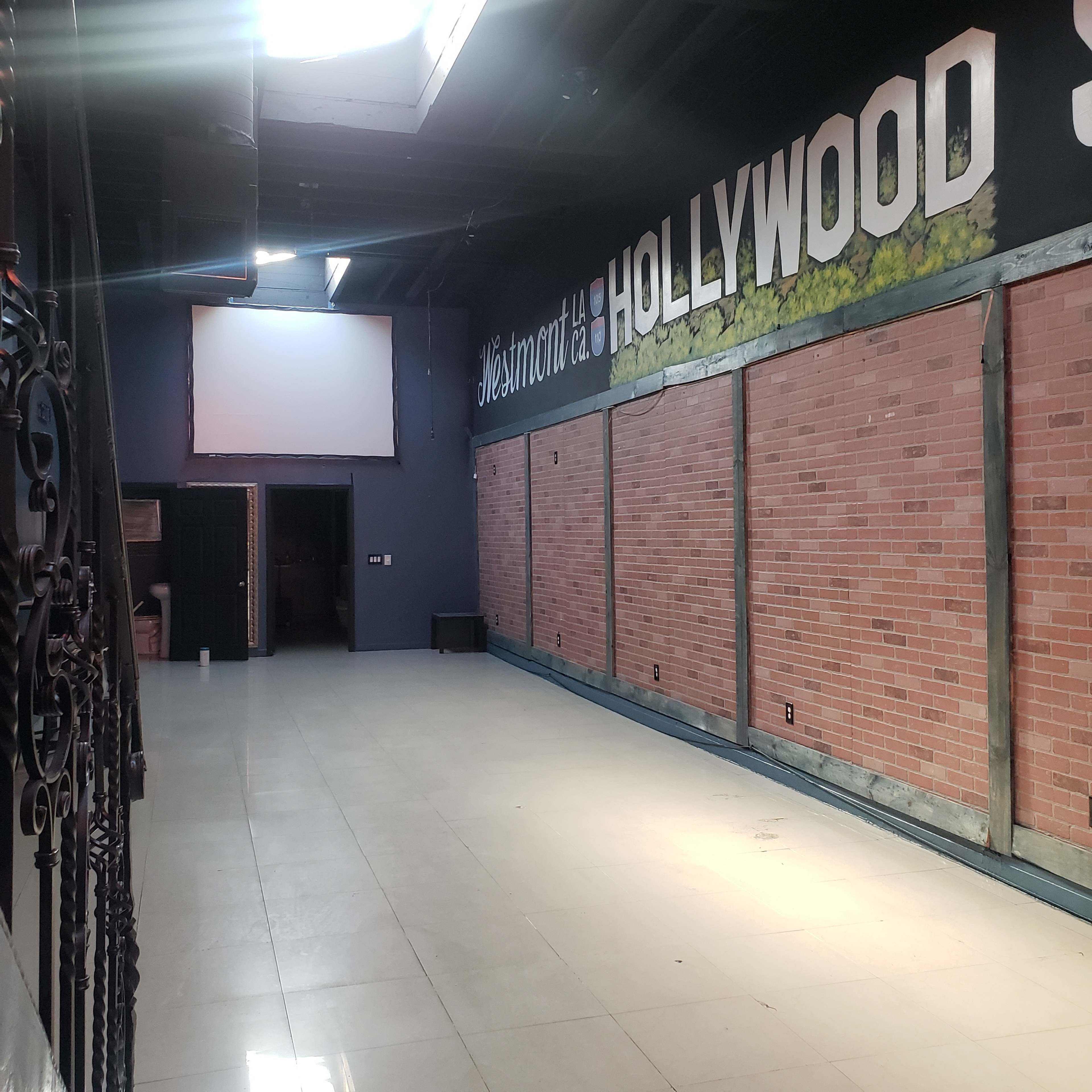 The image shows an empty room with brick walls and a large "Hollywood" mural, featuring a screen at the far end and no furniture visible.