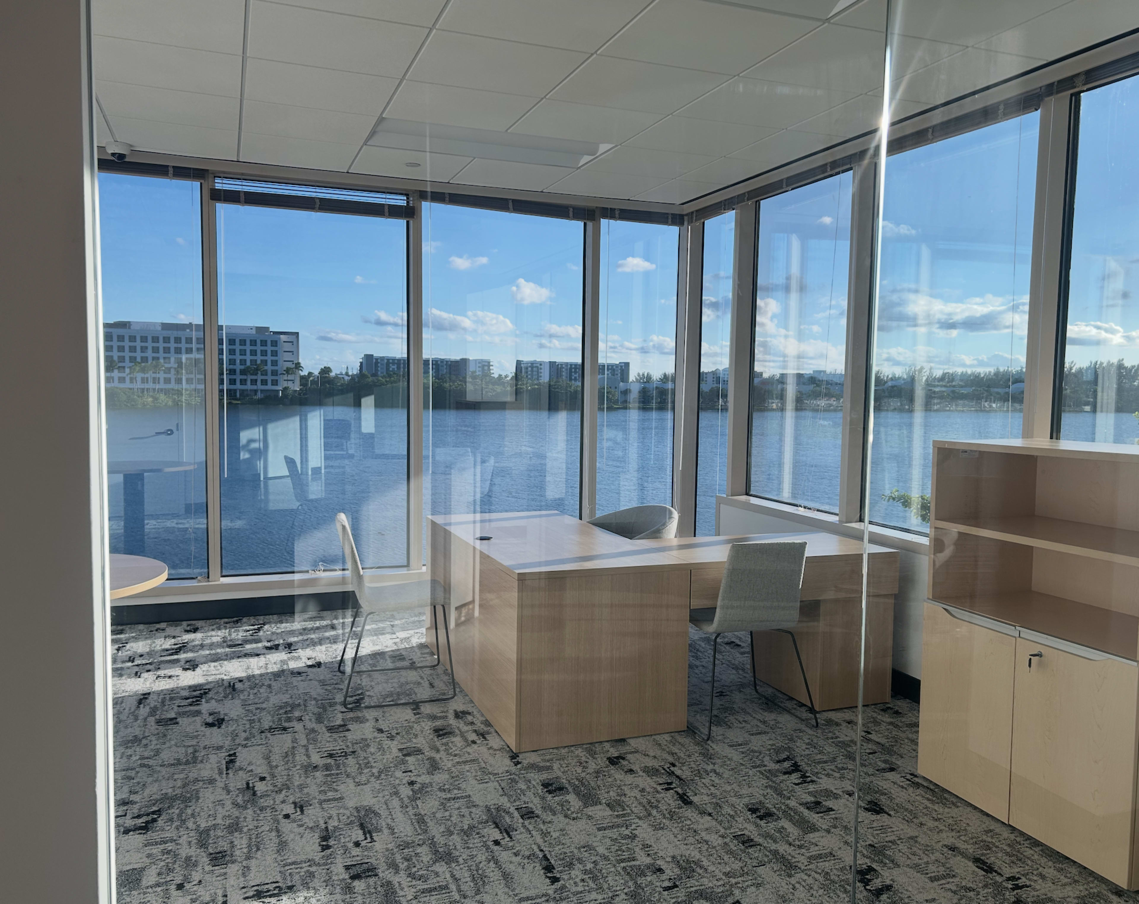 The image shows a modern office space with large windows overlooking a body of water and a view of nearby buildings.