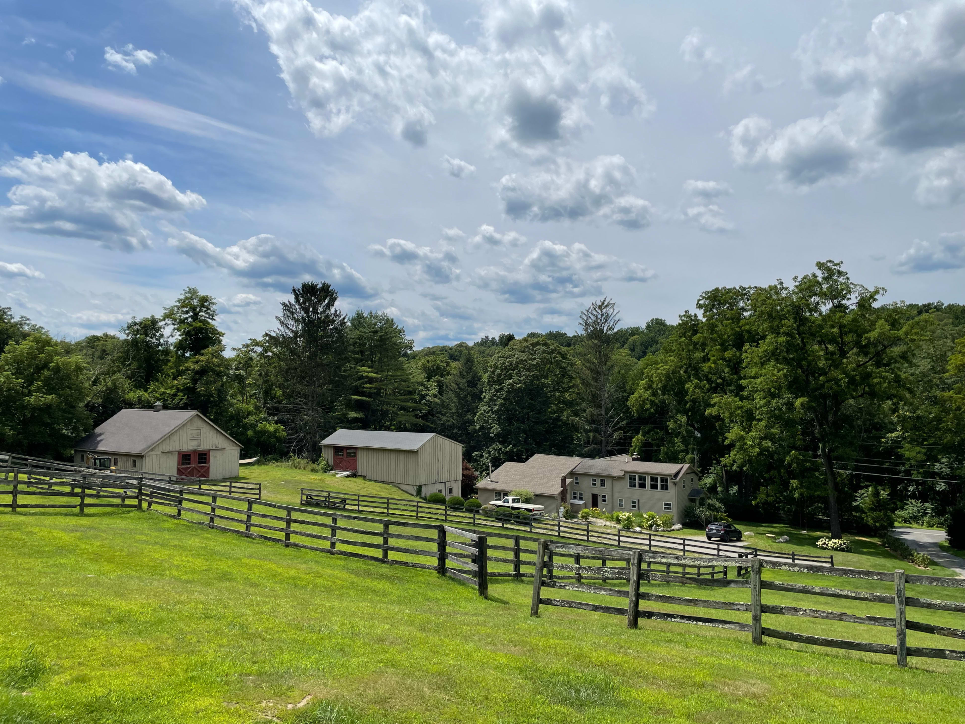 A rural landscape featuring two houses and several outbuildings surrounded by green fields and a wooden fence under a partly cloudy sky.