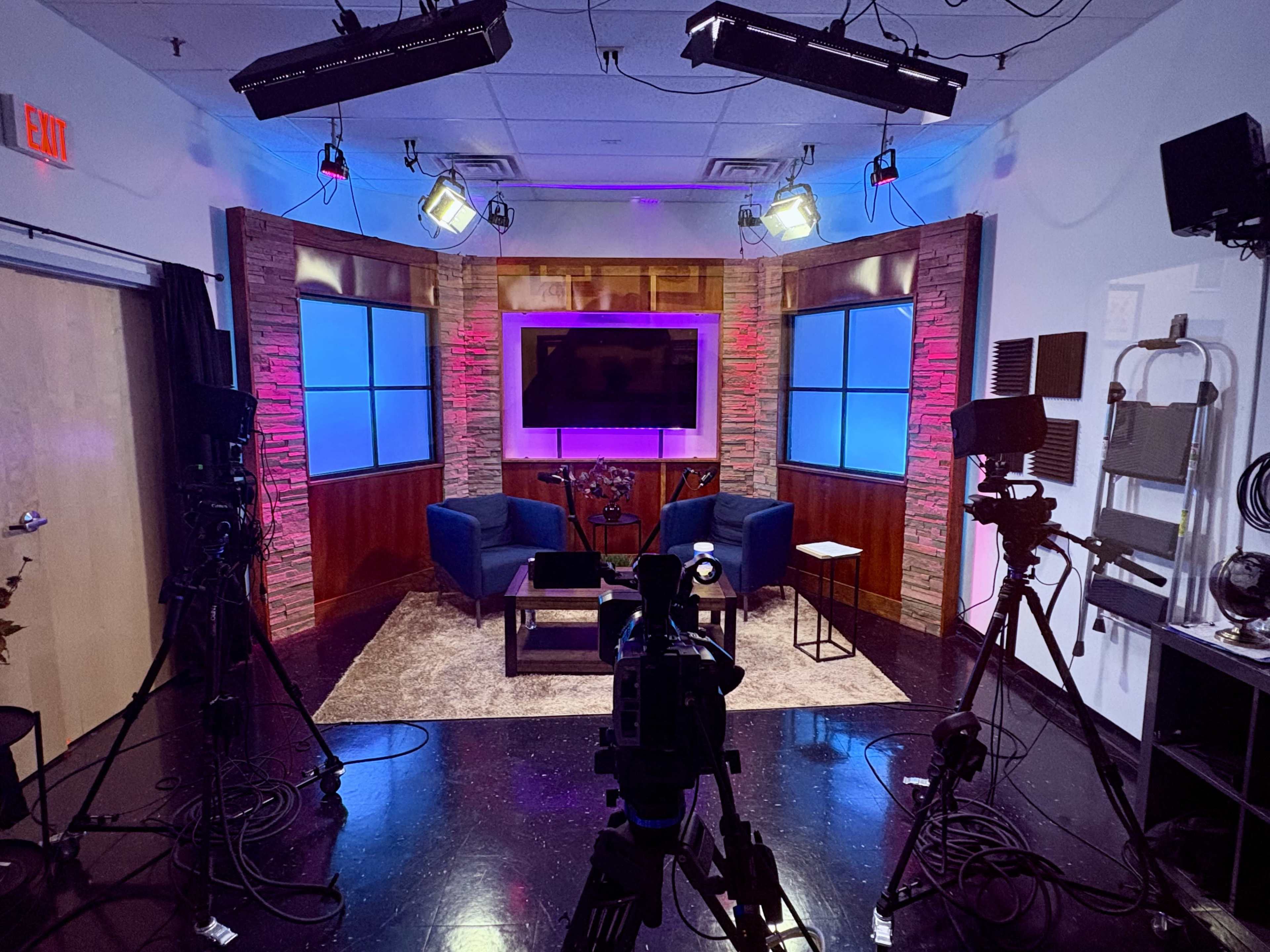 A television studio is set up with two blue chairs, a small table, and multiple cameras positioned around, illuminated by various lights.