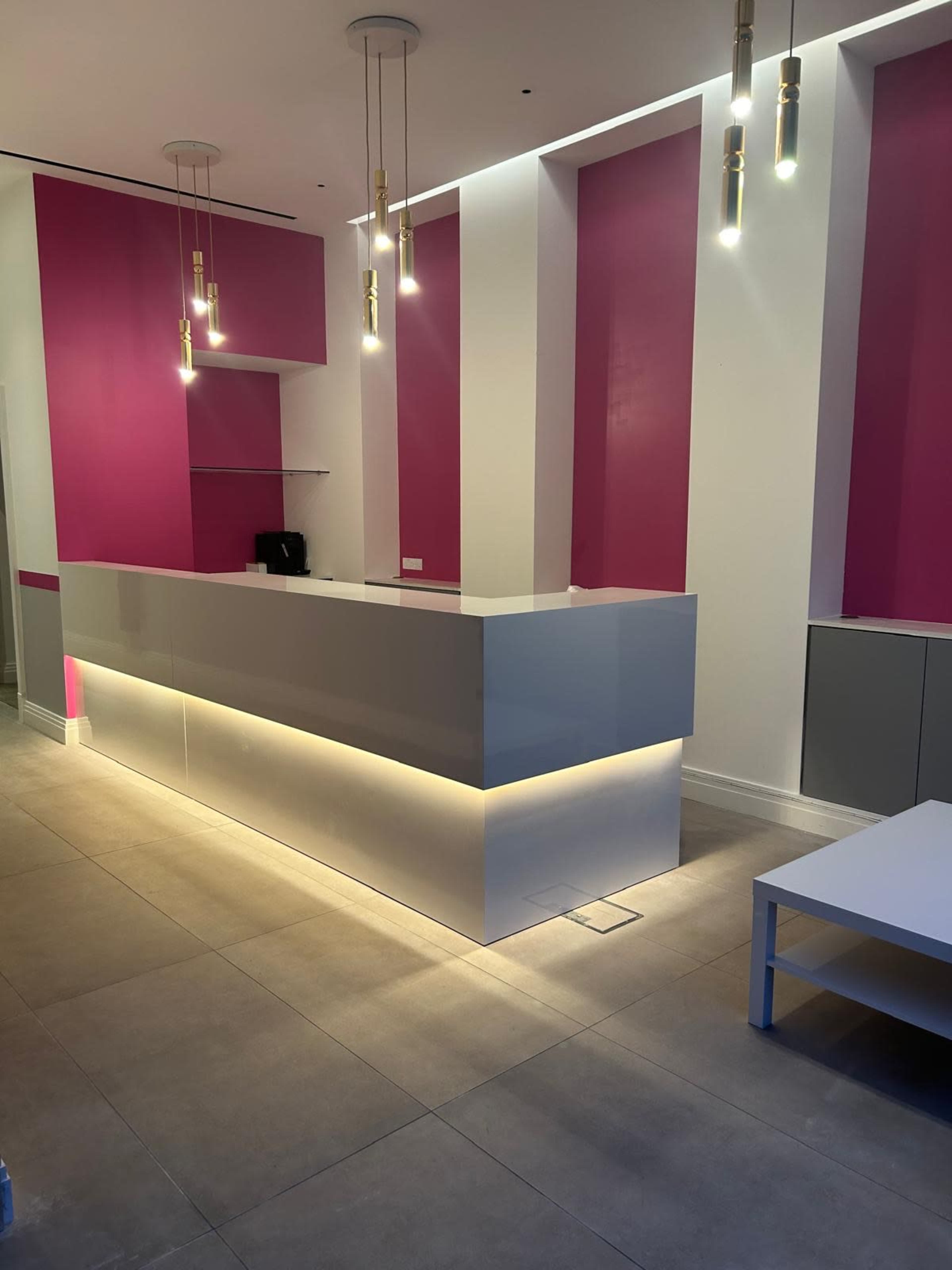 A modern reception area with a sleek gray front desk and pink accent walls, illuminated by pendant lights.