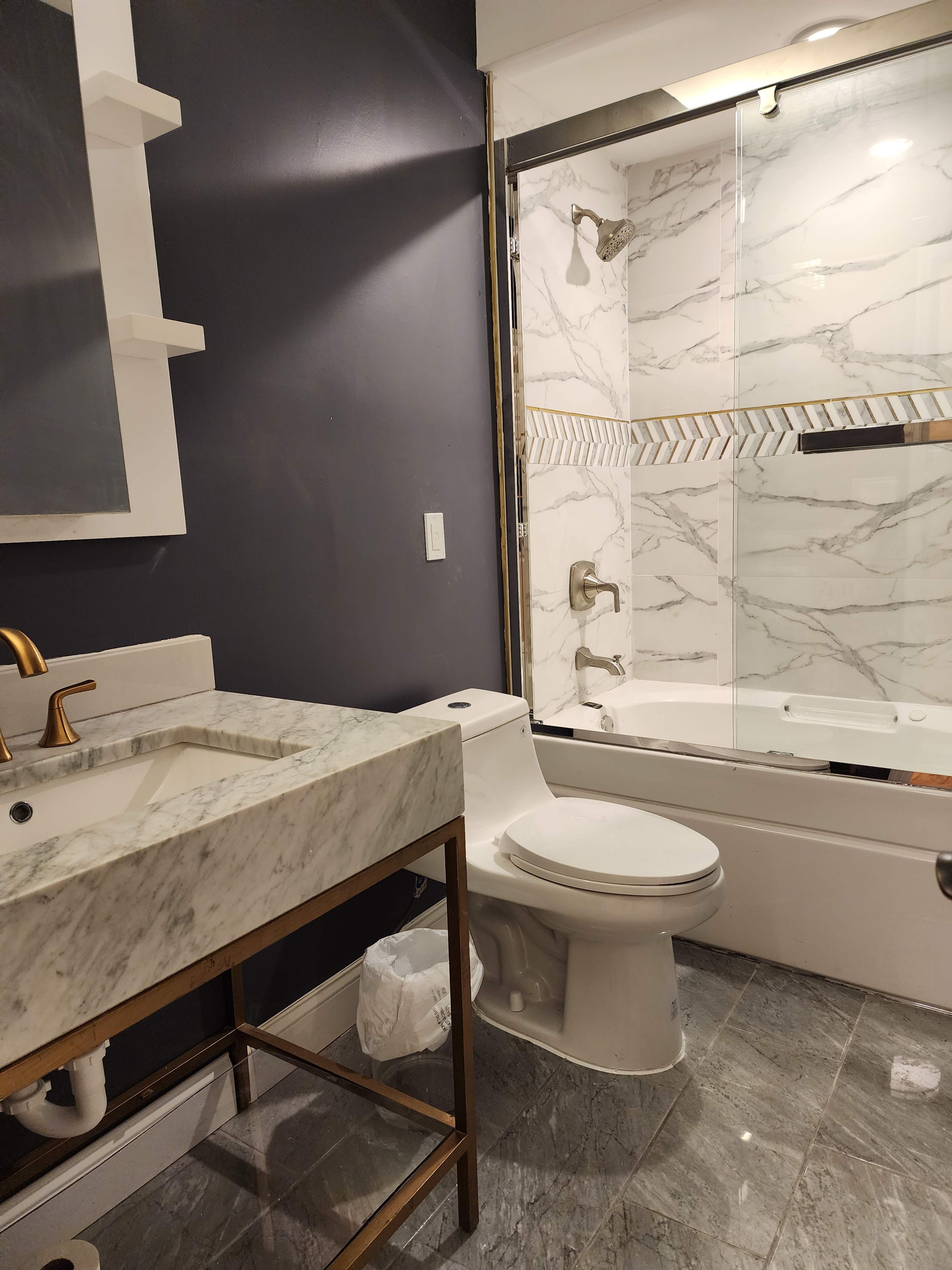 A modern bathroom featuring a marble vanity with a gold faucet, a toilet, and a glass-enclosed shower with marble-patterned tiles.