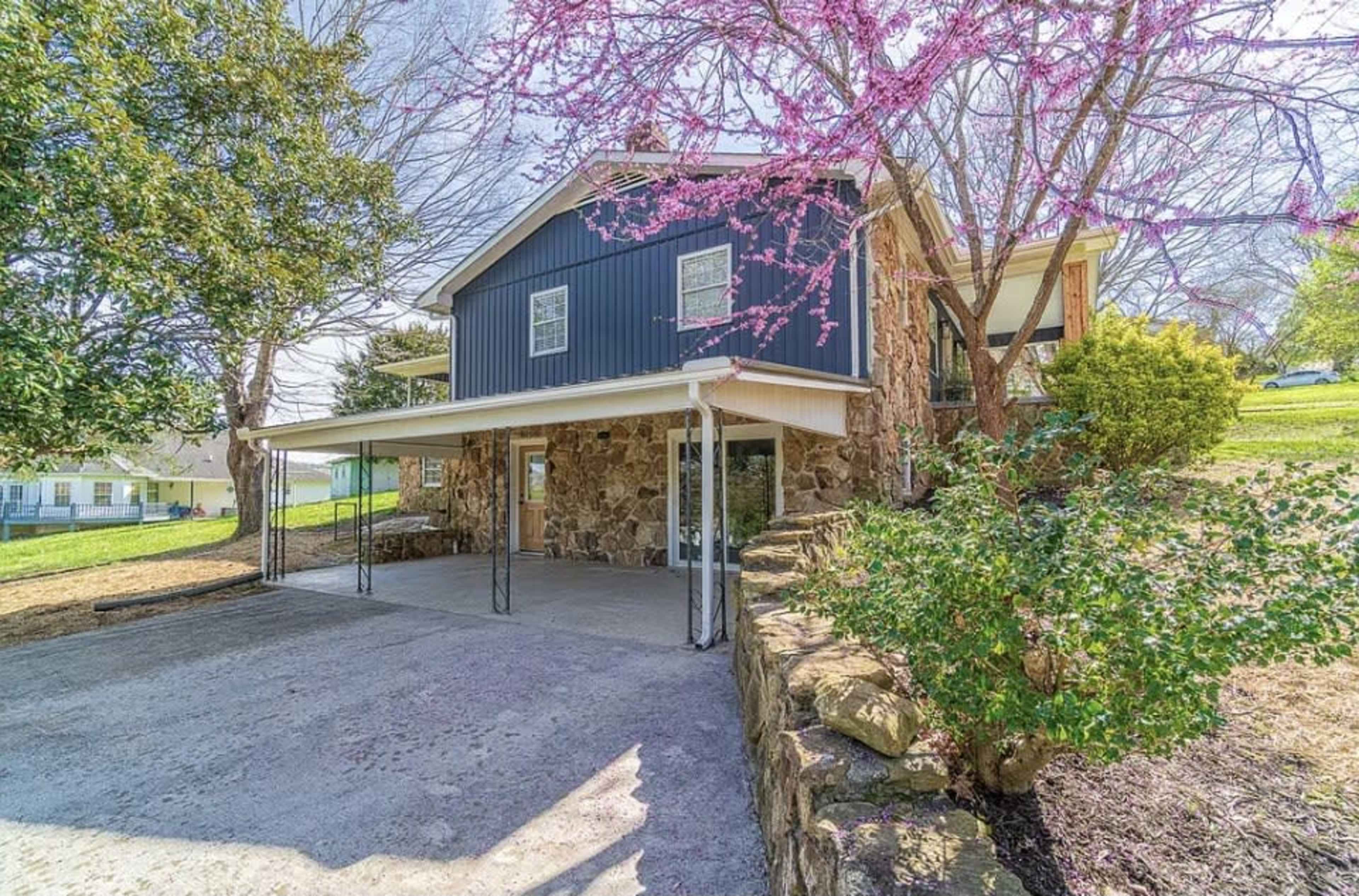 A two-story house with a stone exterior and a covered parking area is surrounded by greenery and a blooming tree.