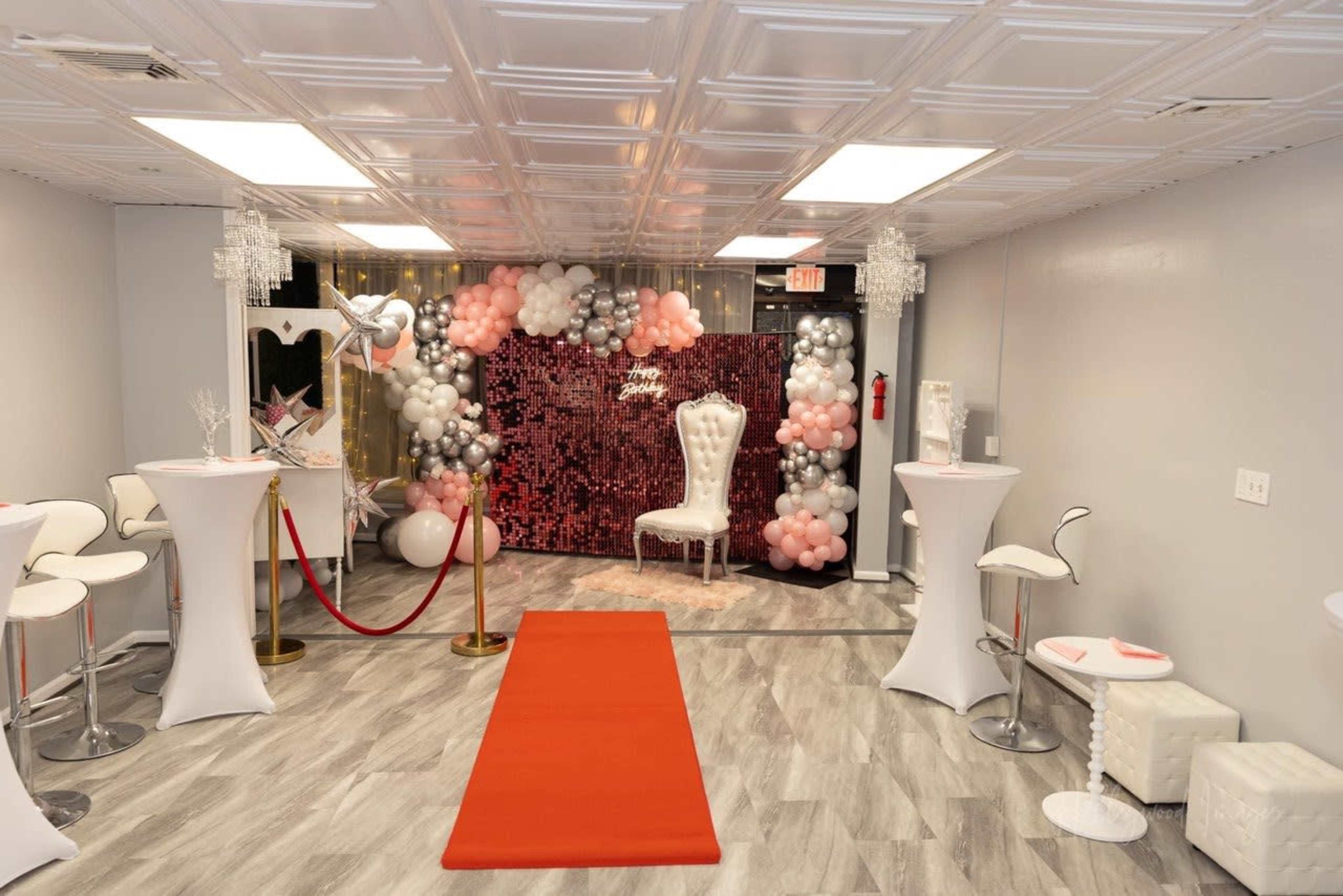 A decorated event space featuring a red carpet leading to a backdrop of balloons and sequins, with white seating and tables arranged around the area.