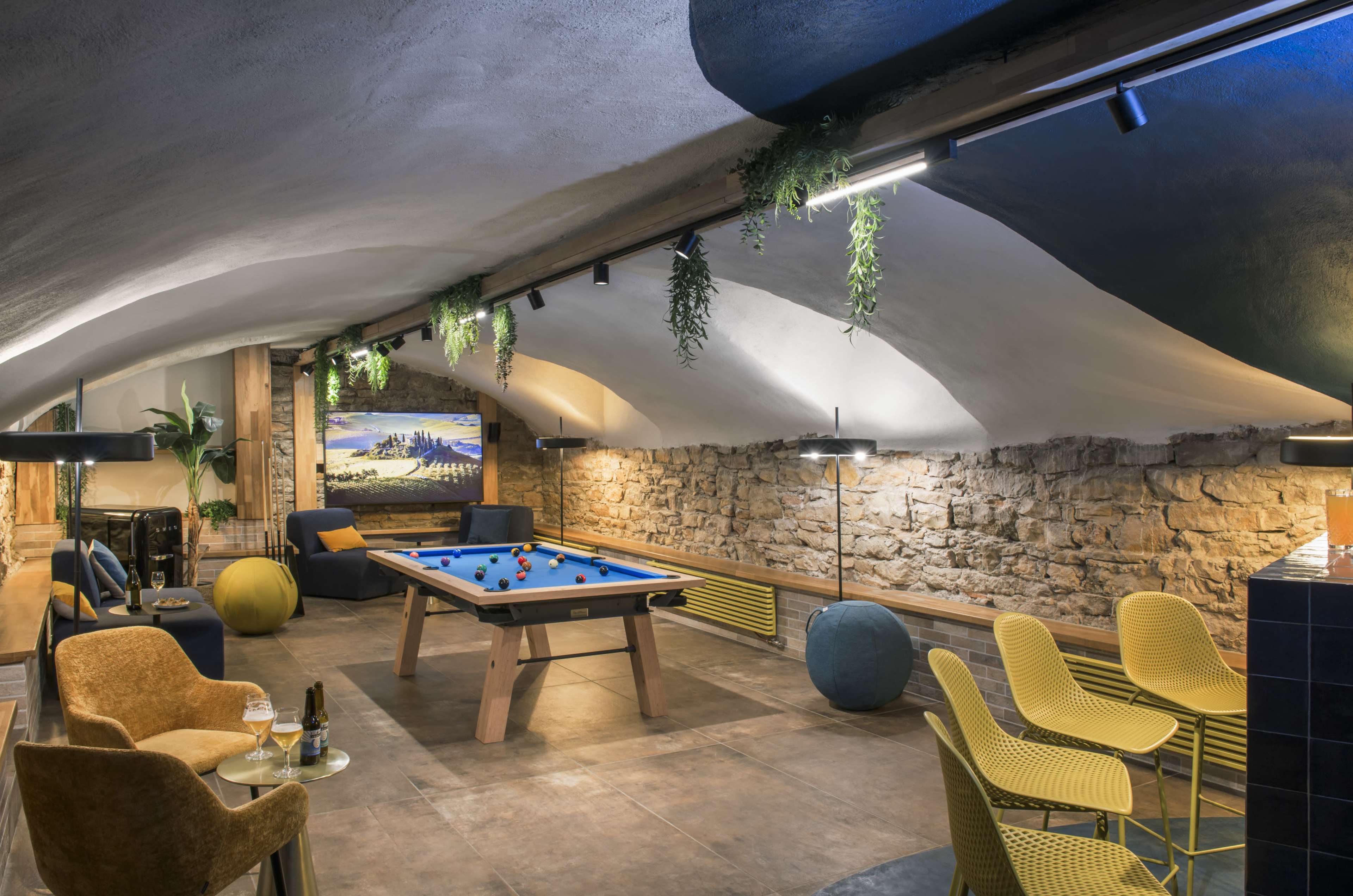 The image shows a stylish game room with a blue pool table, modern seating, and a stone wall under a curved ceiling.