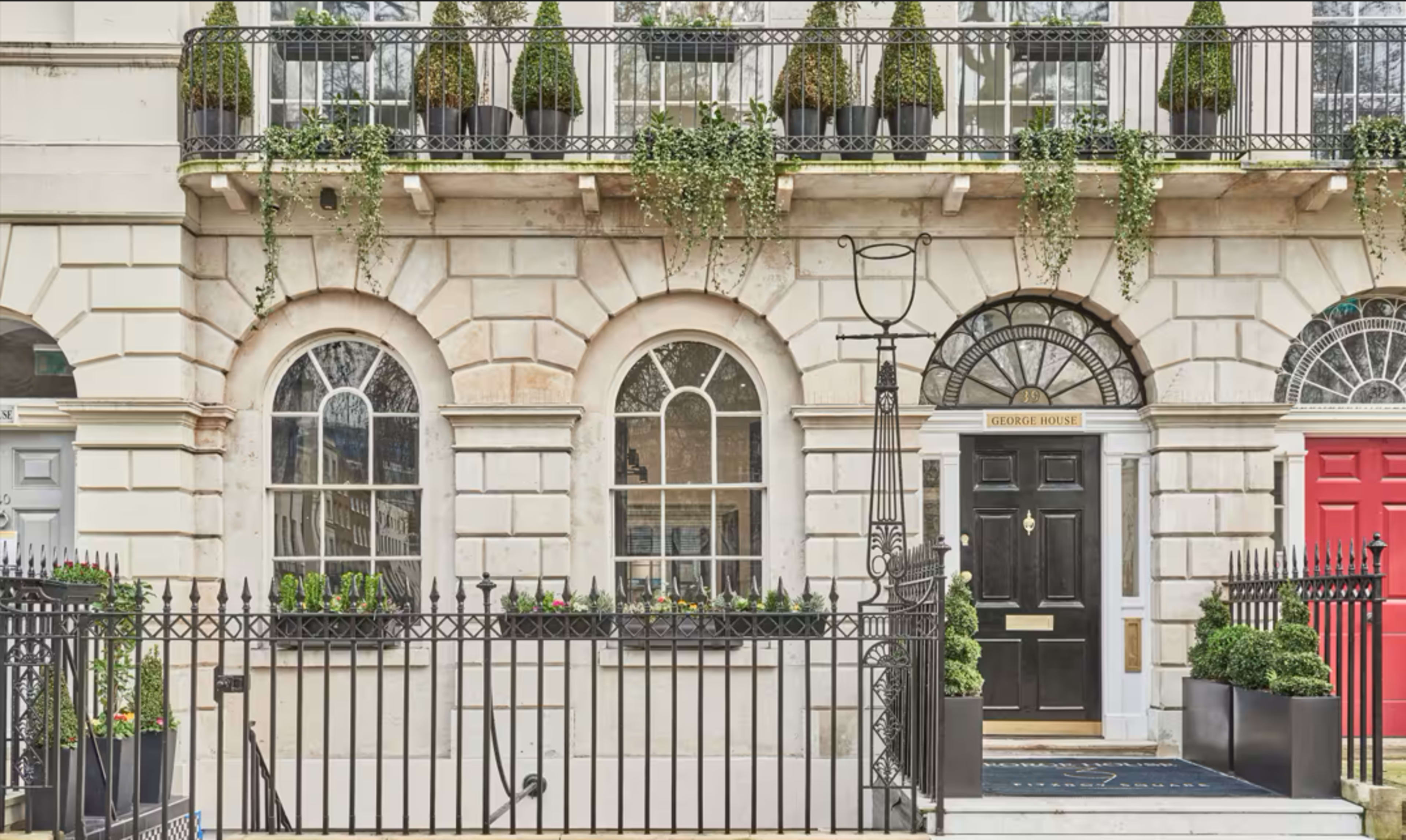 The image shows a classic townhouse facade with arched windows, a black front door, and a gated entrance adorned with potted plants.