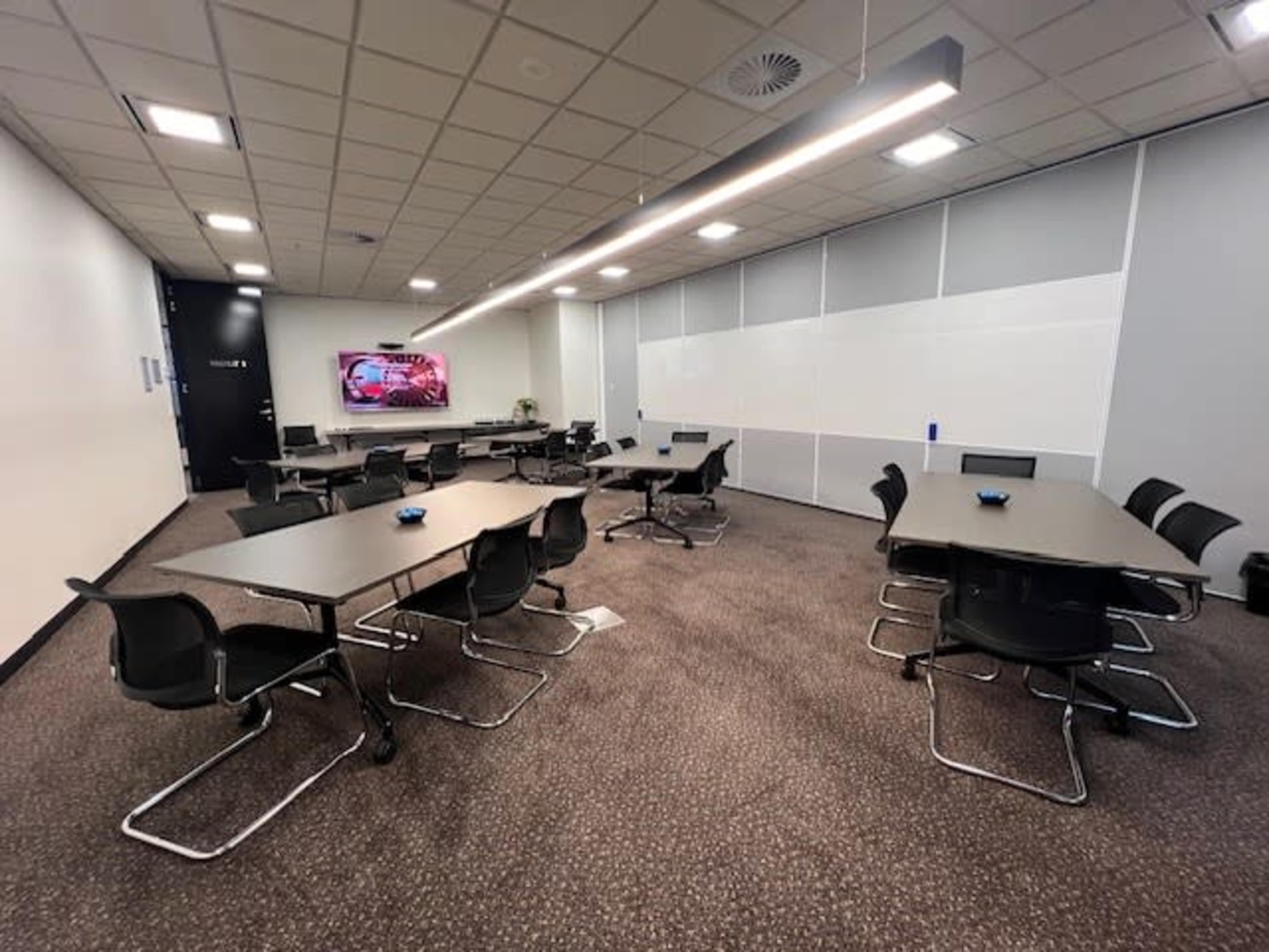 The image shows a modern conference room equipped with multiple tables and chairs, a wall-mounted display, and a whiteboard.
