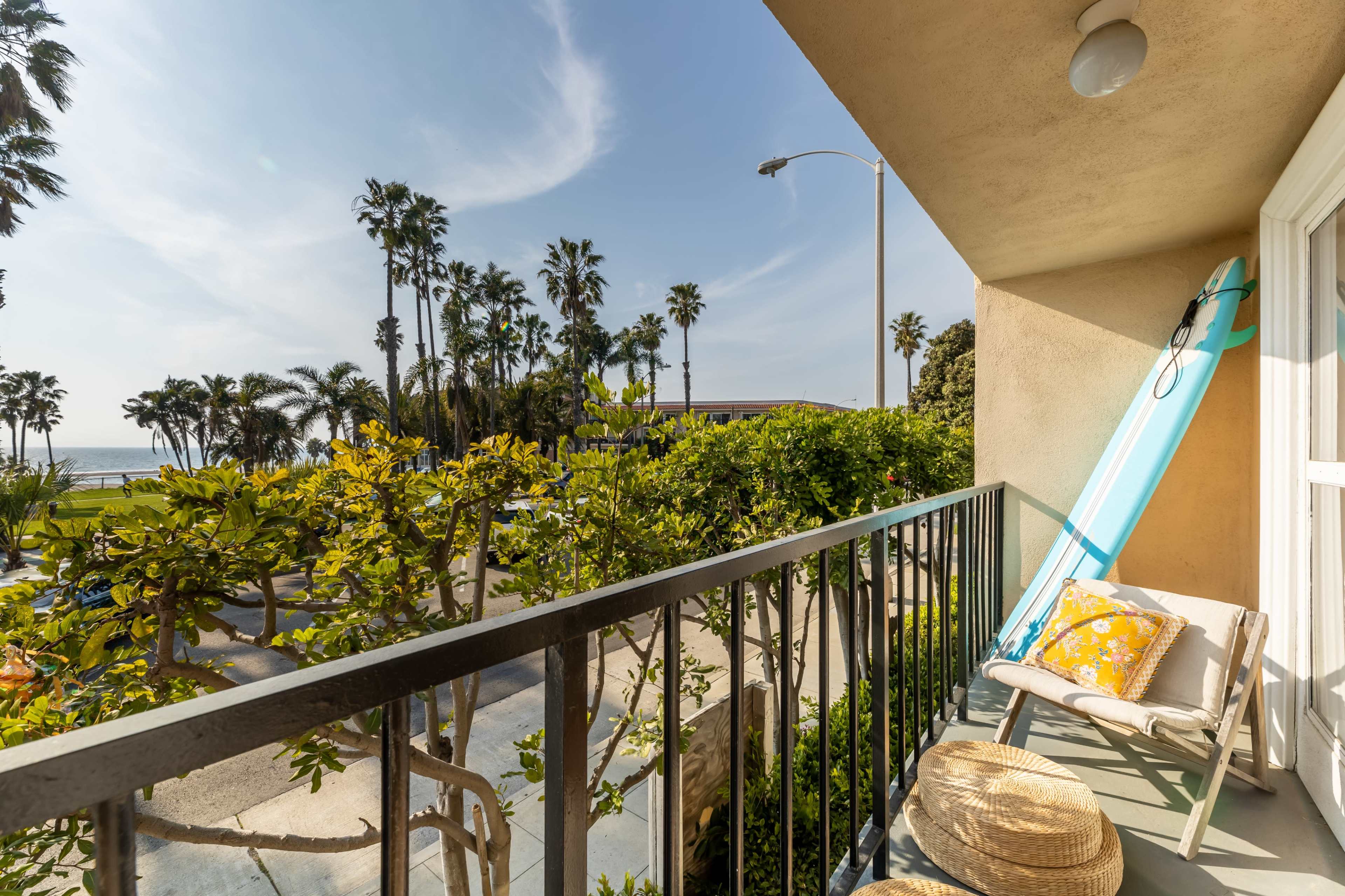 A balcony features a surfboard leaning against the wall, overlooking palm trees and the ocean in the background.