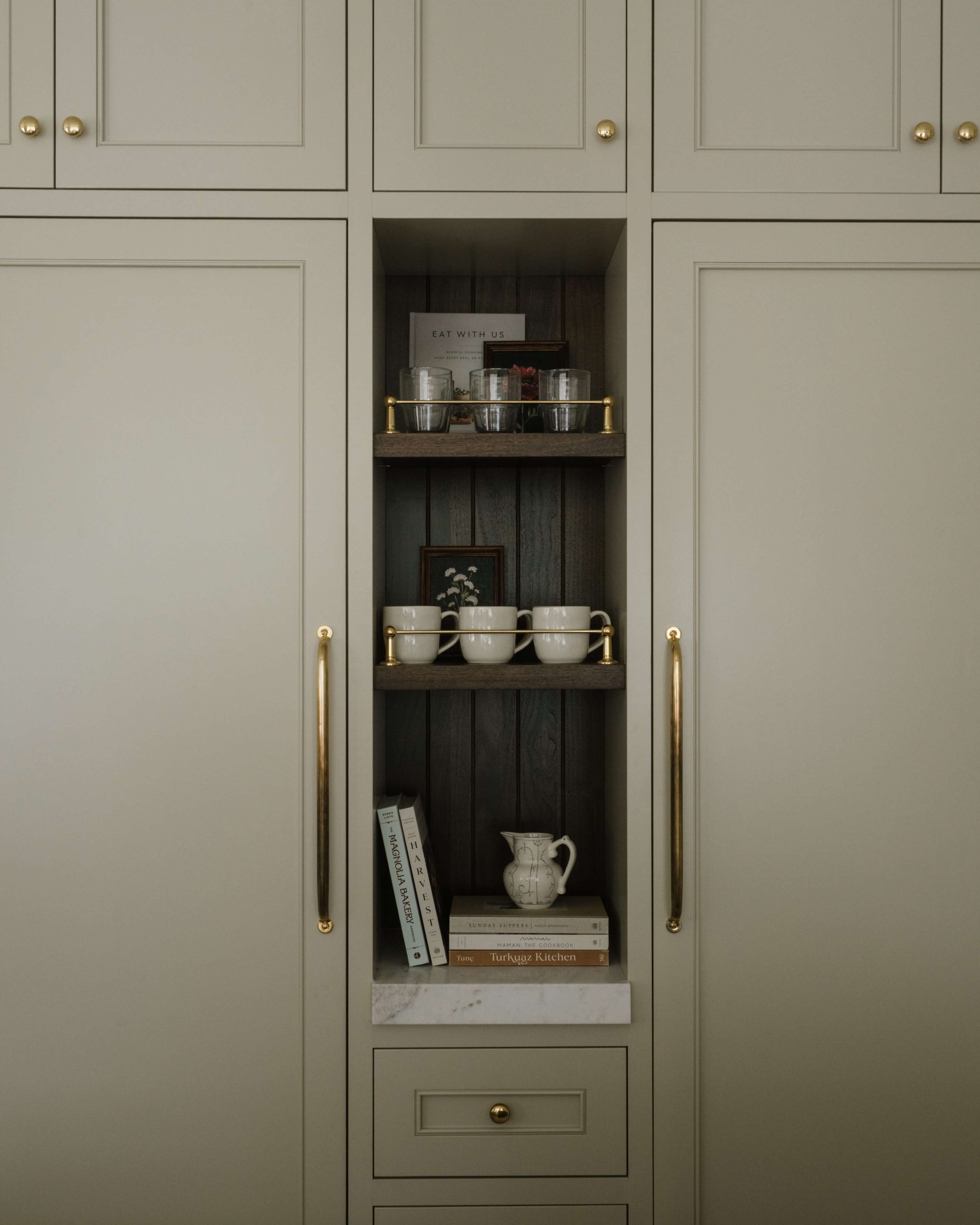 The image features a narrow, built-in shelving unit with glassware and decorative items arranged on wooden shelves, flanked by cabinet doors in a light color.