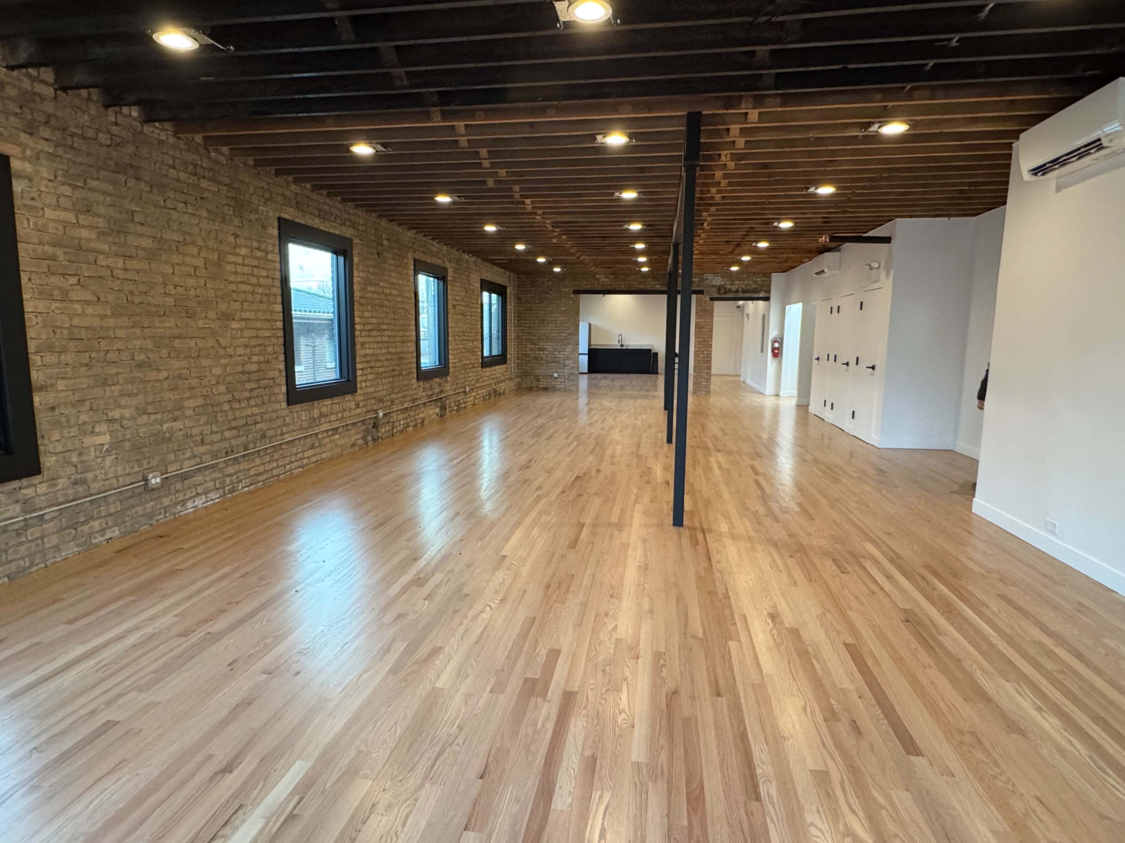 The image shows an empty, spacious room with wooden flooring, exposed brick walls, large windows, and overhead lighting.