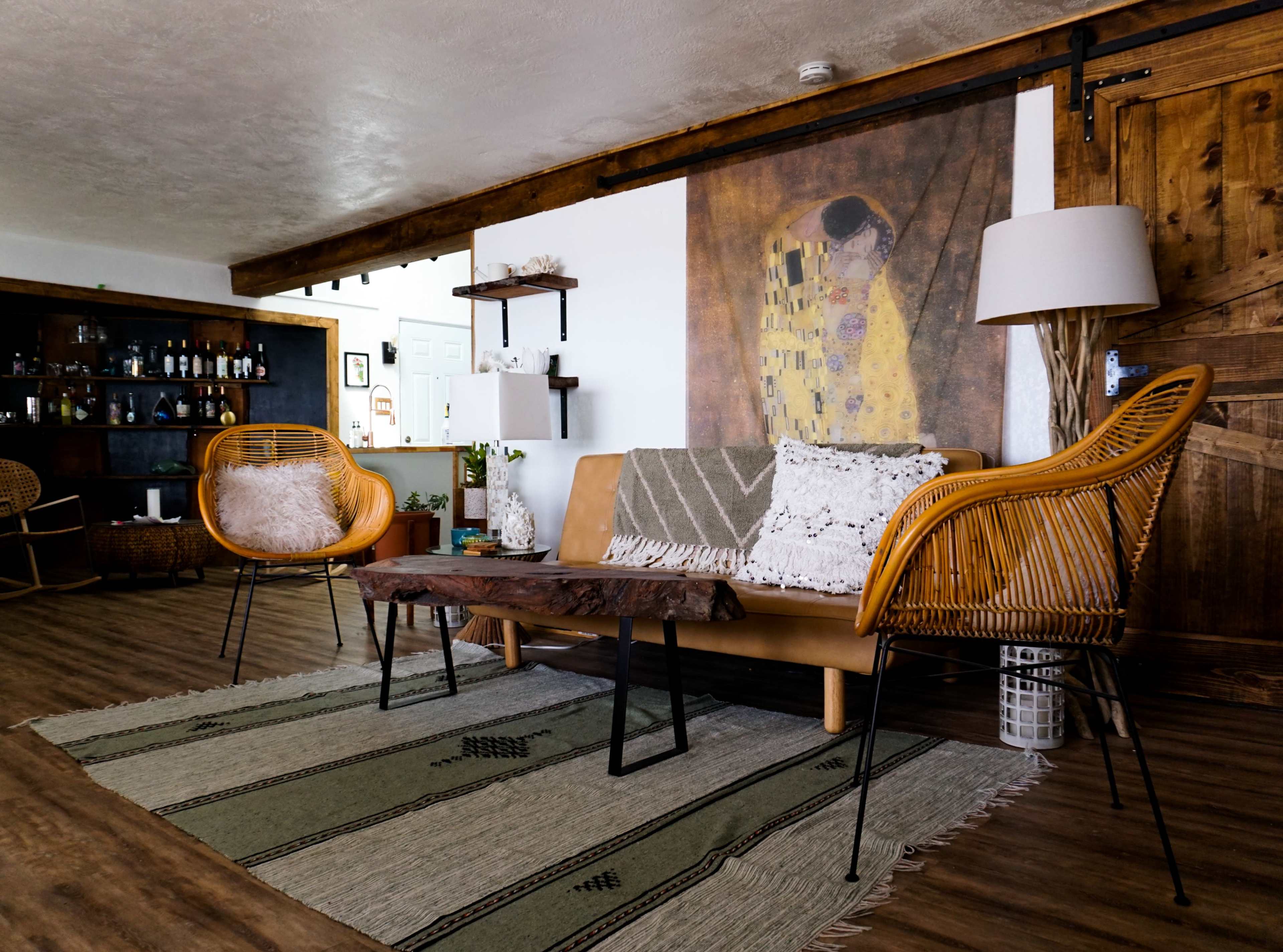 A cozy living area featuring a brown leather sofa, wicker chairs, a wooden coffee table, and a patterned rug, with a large artwork on the wall and a bar area in the background.
