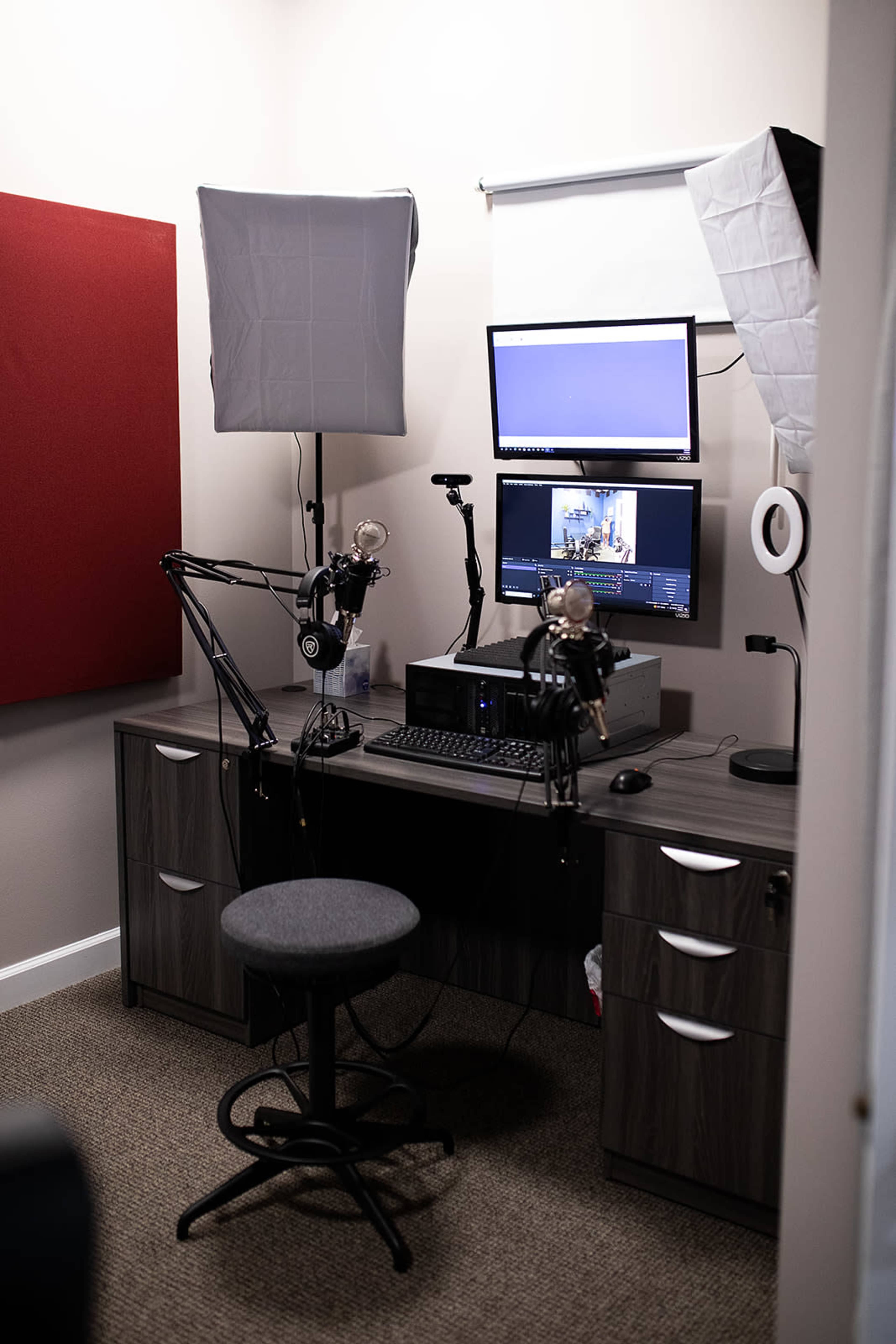 A recording setup featuring a desk with a computer monitor, a microphone on adjustable arms, softbox lights, and a swivel stool in a small room.