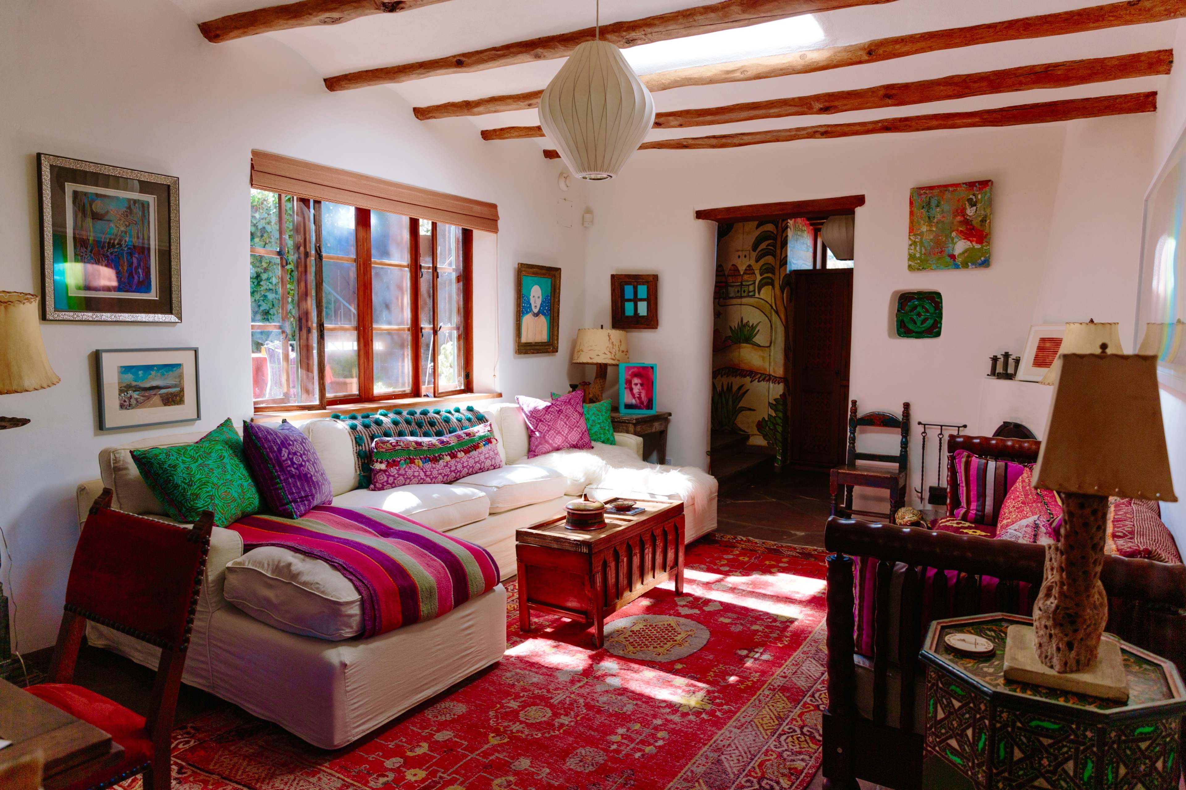 A brightly lit living room featuring a white sofa adorned with colorful cushions, a wooden coffee table, and various artworks on the walls.