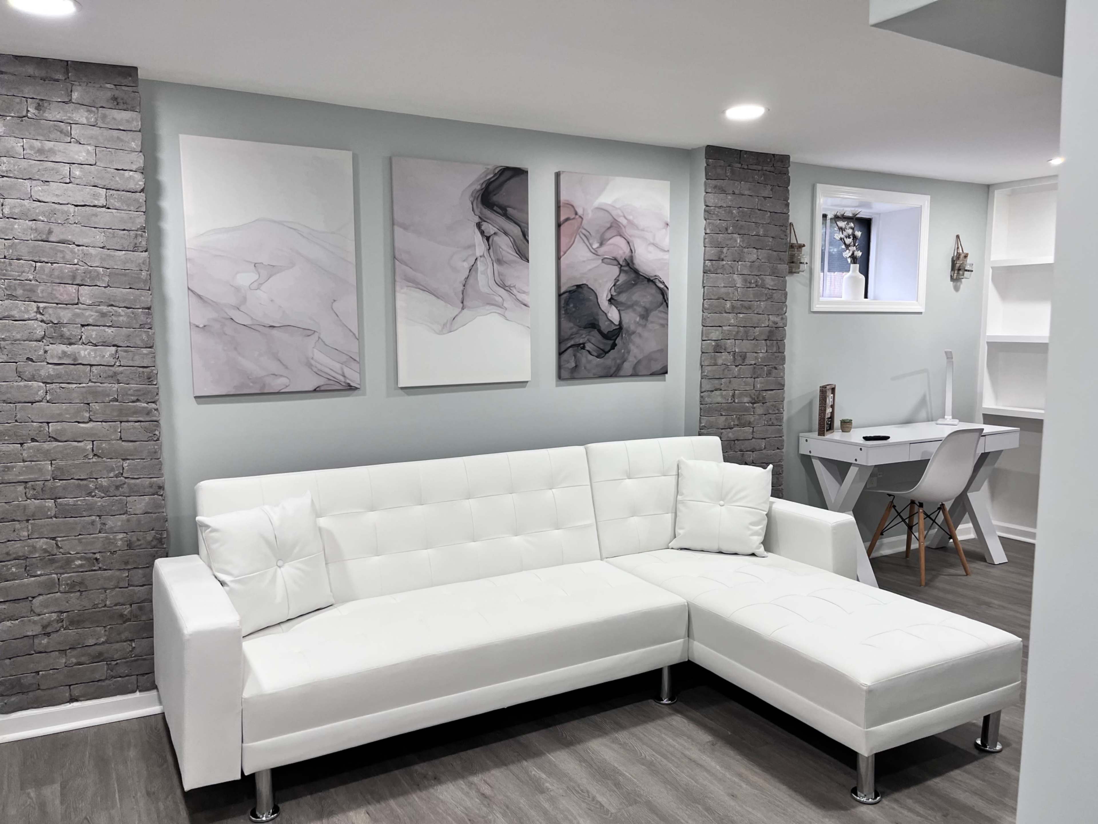 A modern living space features a white sectional sofa against a gray brick wall, complemented by three abstract artworks and a small desk area.