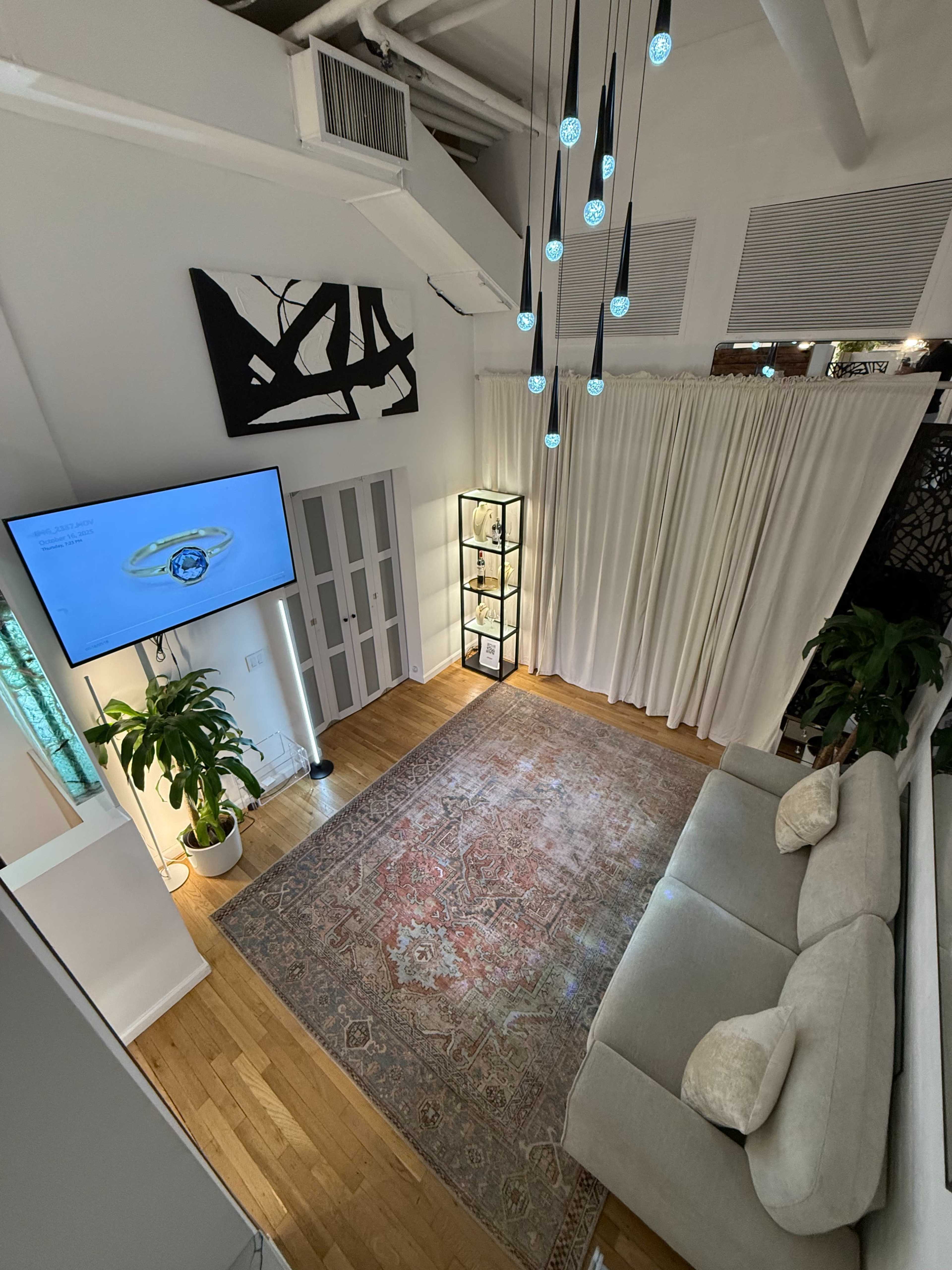 The image shows a modern, well-lit room featuring a gray sofa, a television mounted on the wall, a display cabinet with glass doors, and pendant lights hanging from the ceiling.