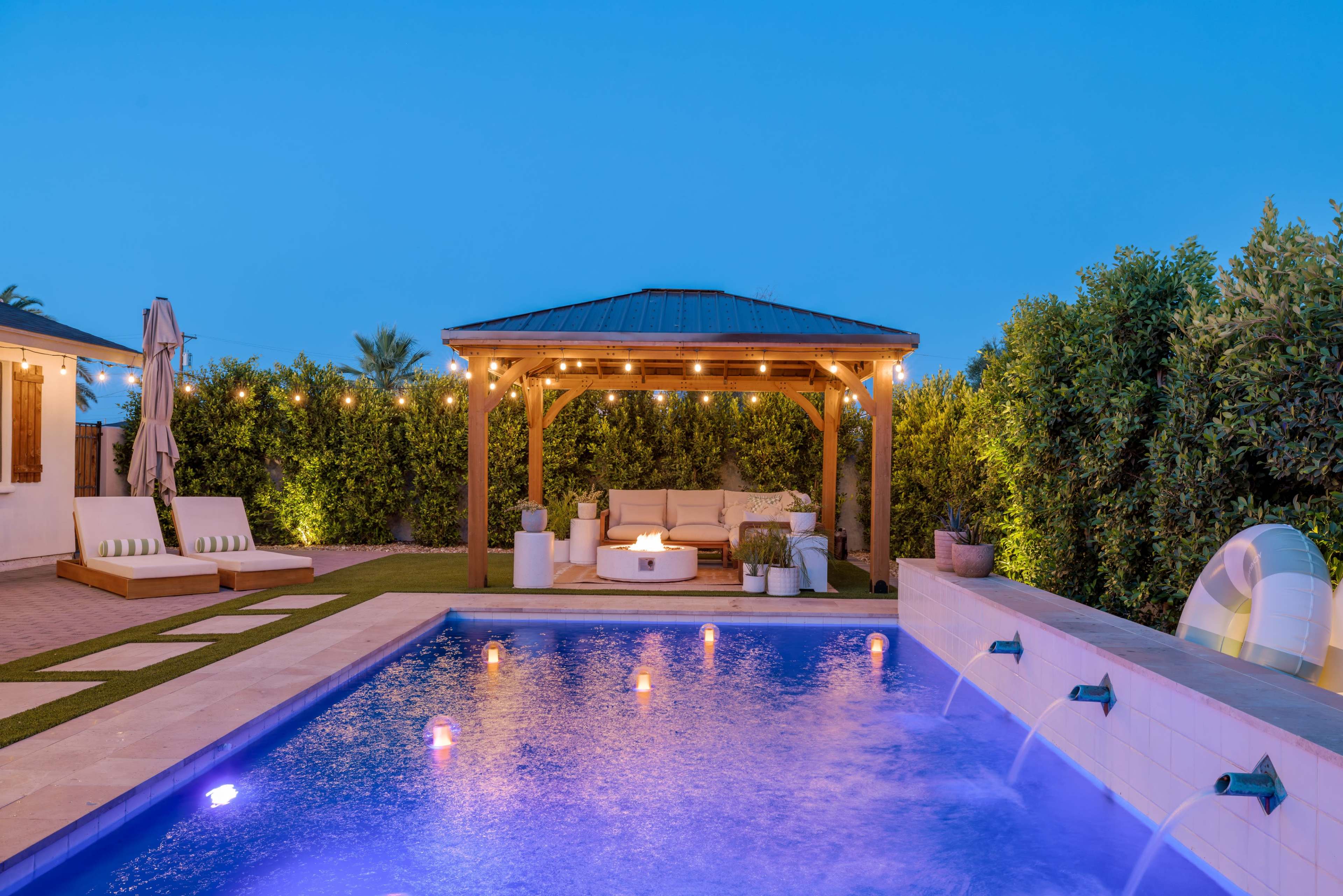 The image shows a well-lit backyard at dusk, featuring a swimming pool with illuminated water features, a covered seating area, and lounge chairs surrounded by greenery.