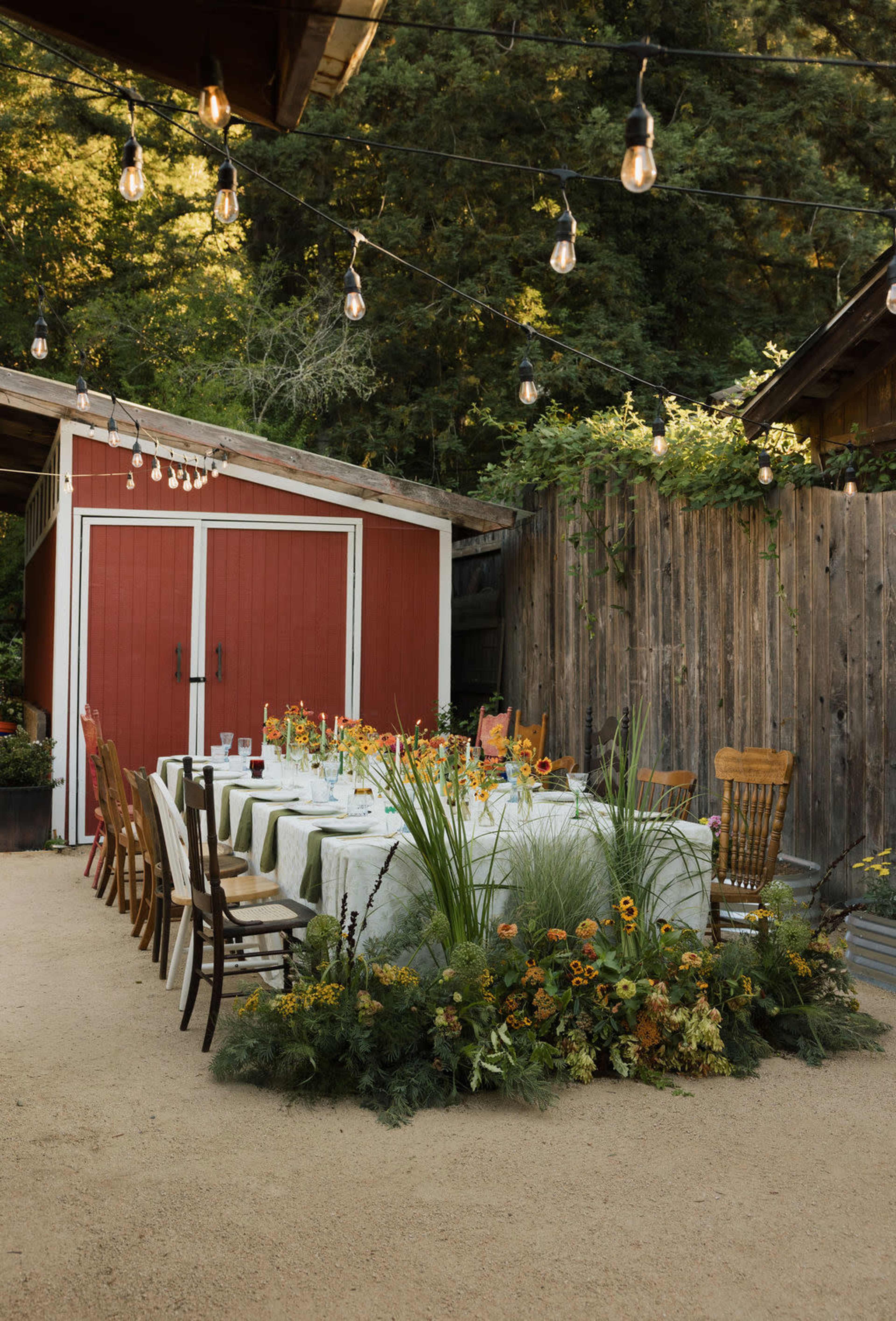 A long dining table is set outside near a red shed, surrounded by string lights and decorated with flowers and greenery.