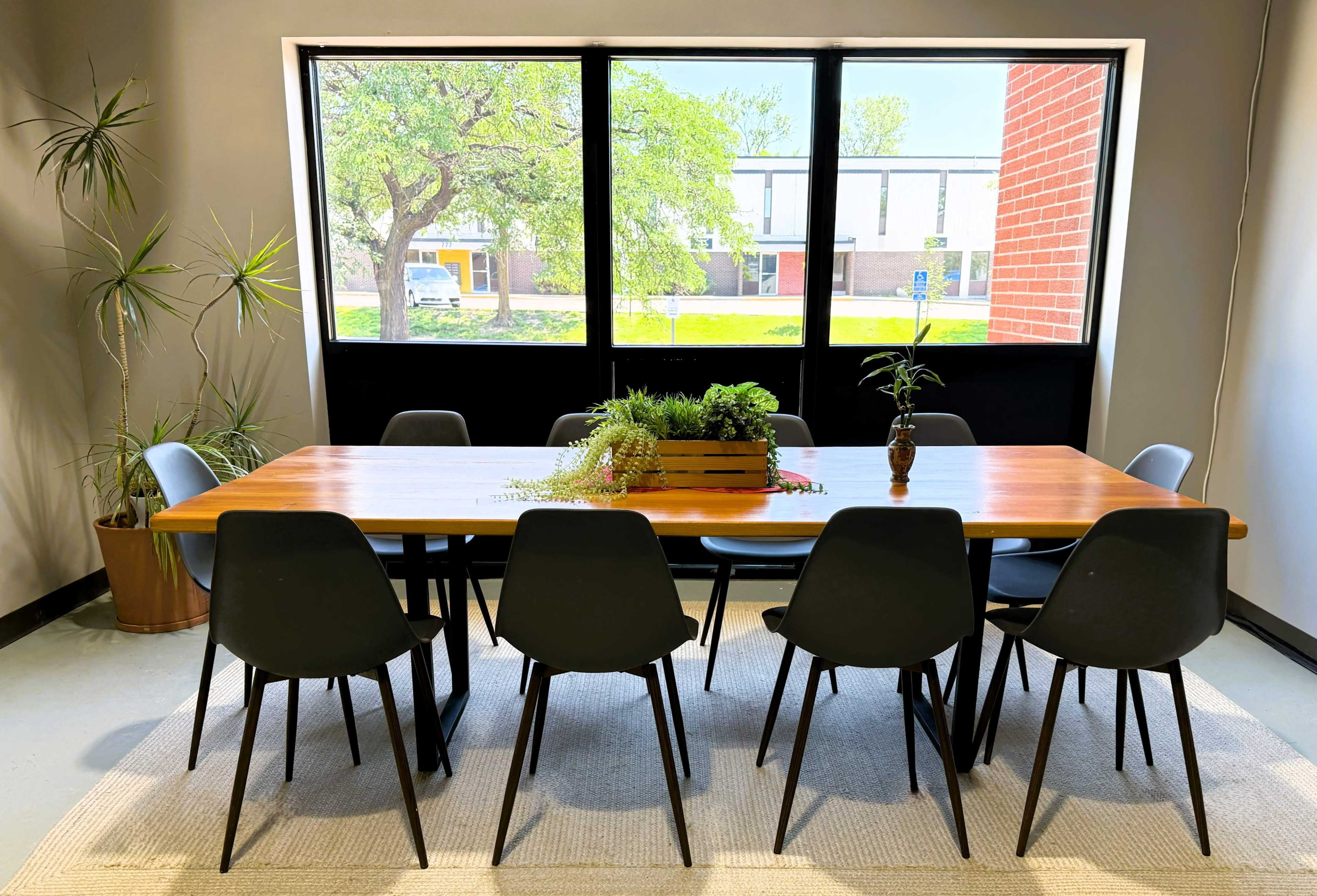 A long wooden table with eight black chairs sits in front of large windows, with plants on the table and in the corners of the room.