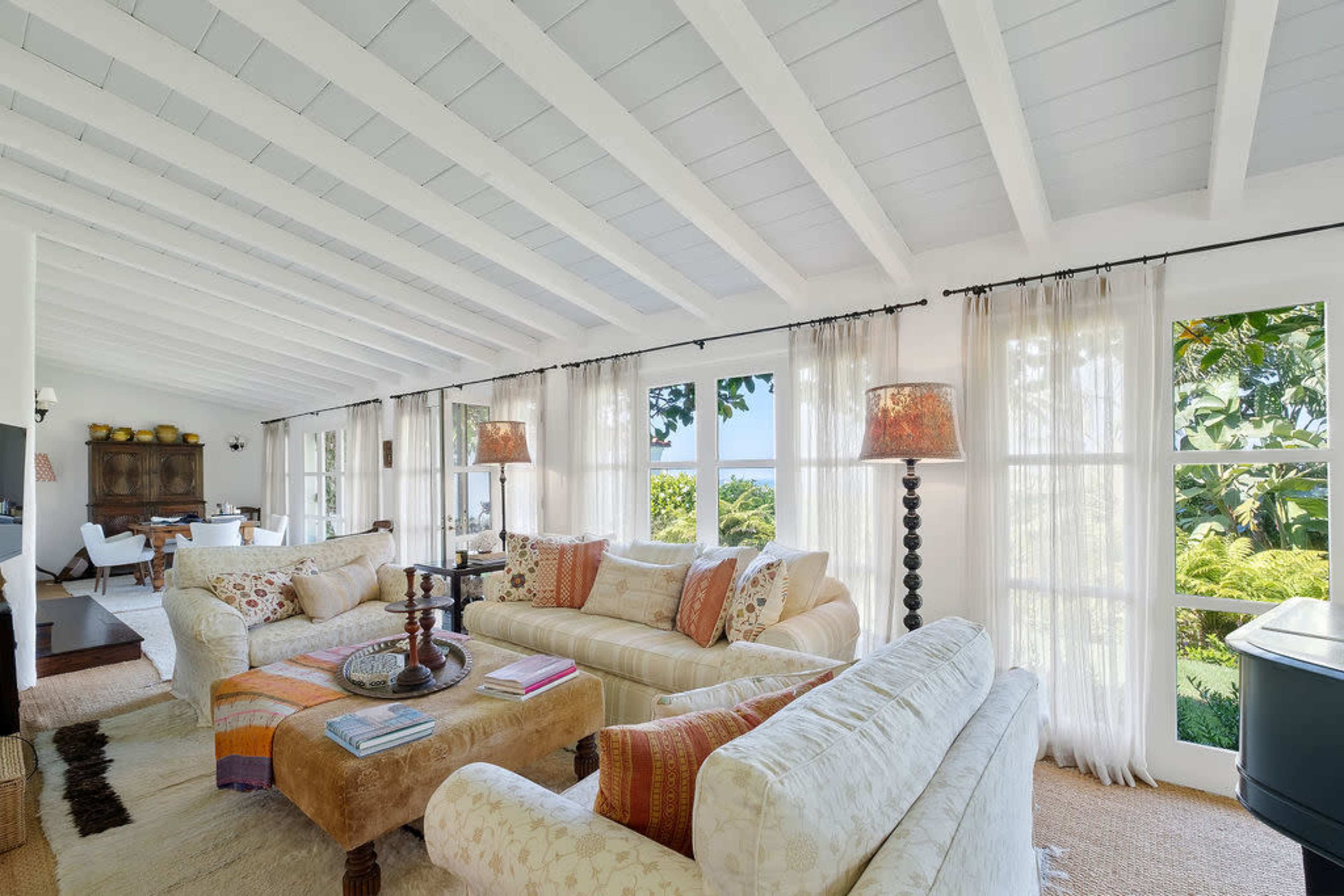 The image shows a bright, airy living room with white beams, large windows, and a mix of sofas and decorative elements overlooking a garden.