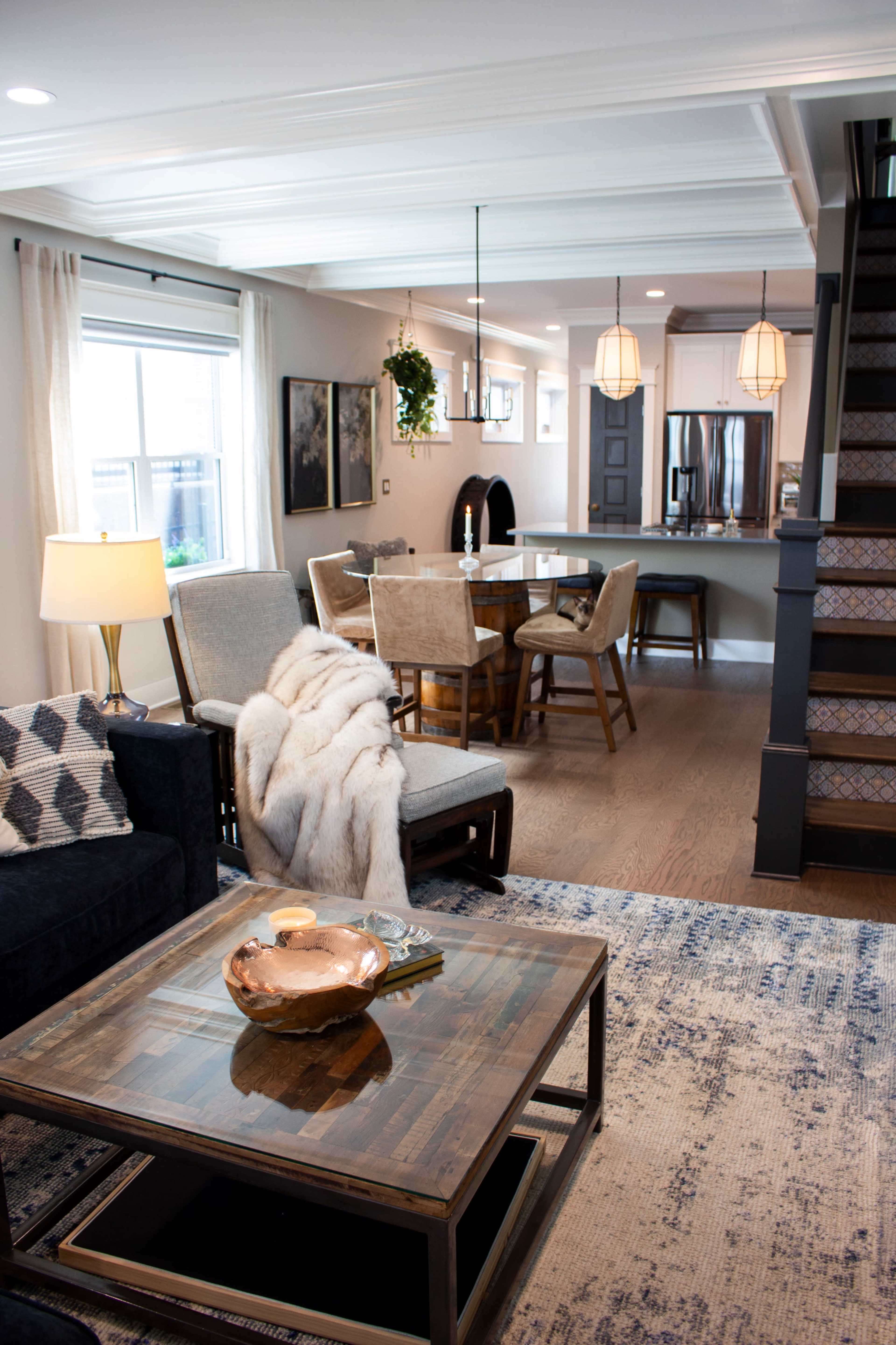 The image shows a modern living room and dining area with a cozy atmosphere, featuring a mix of furniture styles, a dining table set for four, and an open layout leading to a staircase.