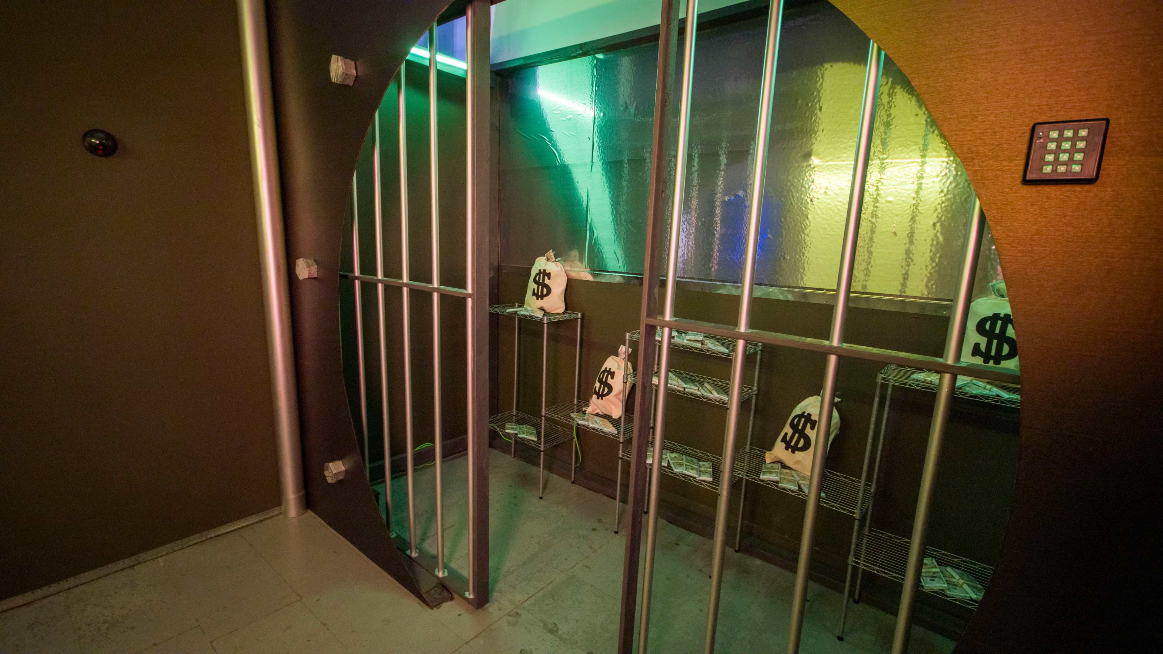 The image shows an enclosed room with metallic bars and bags marked with dollar signs inside wire shelving.