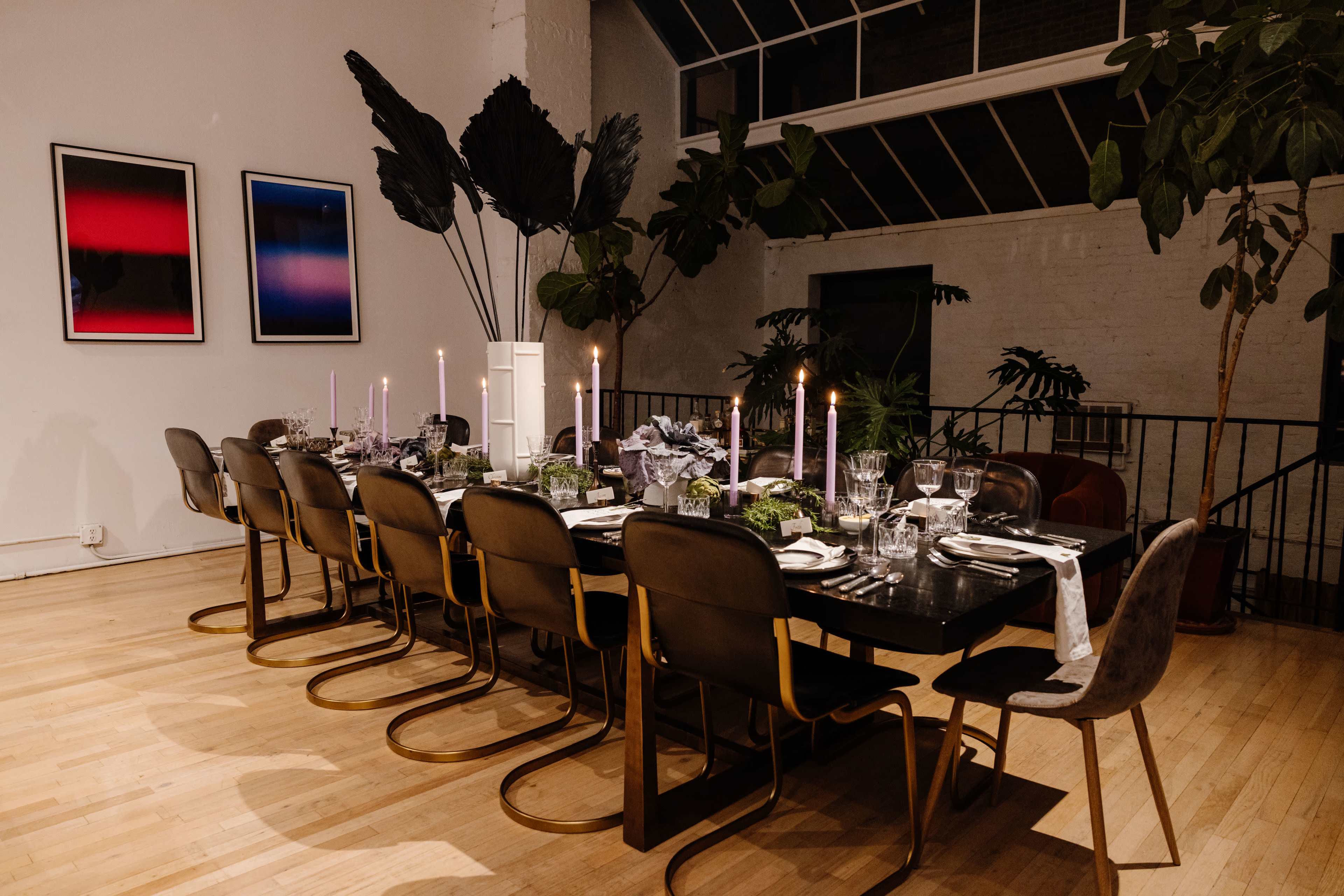 A long dining table is set with candles and tableware in a modern, well-lit room featuring large plants and abstract artwork on the walls.