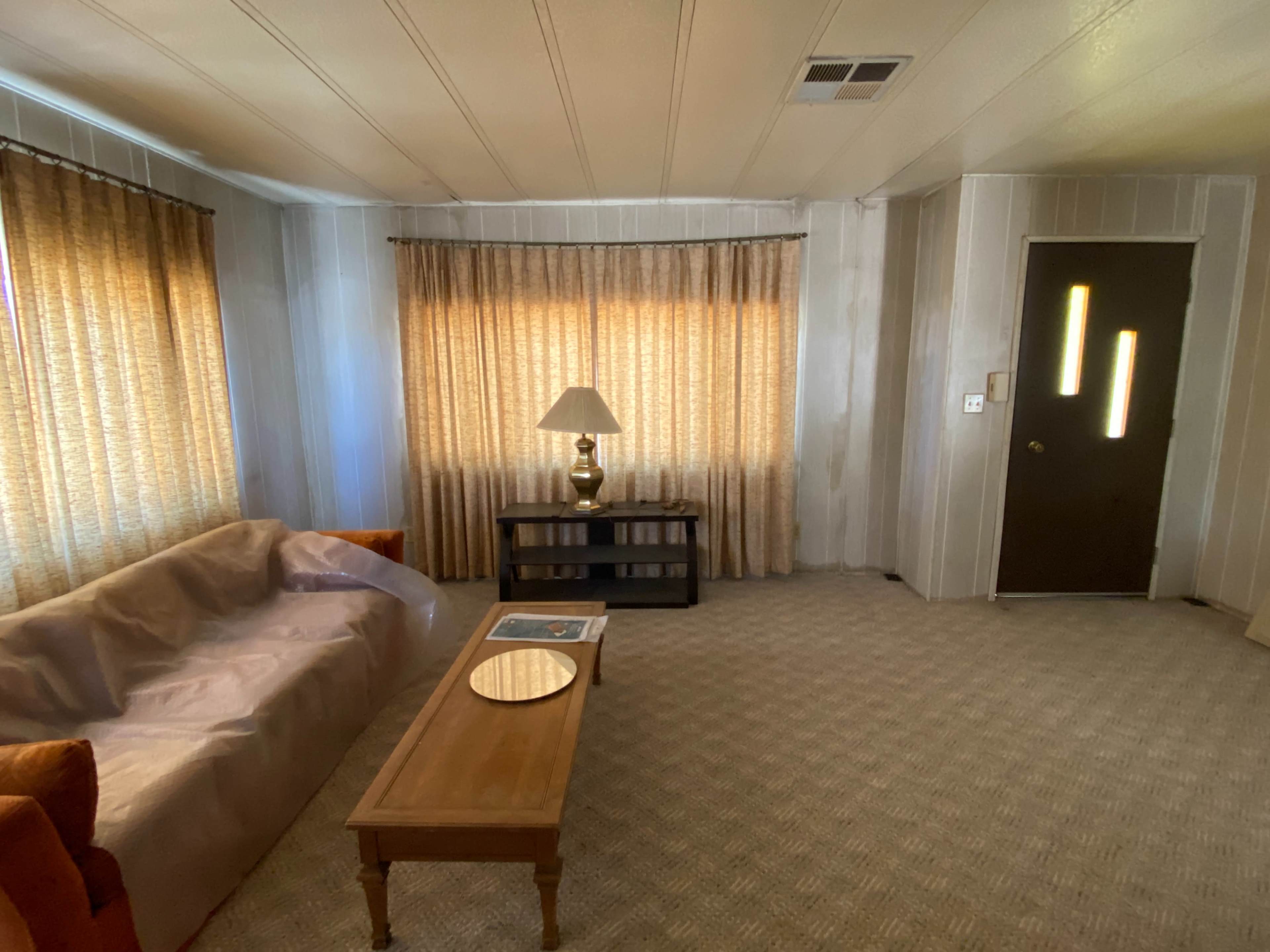 The image depicts a living room with a beige couch, a wooden coffee table, a lamp, and two large curtains letting in warm light, along with a door featuring two vertical windows.
