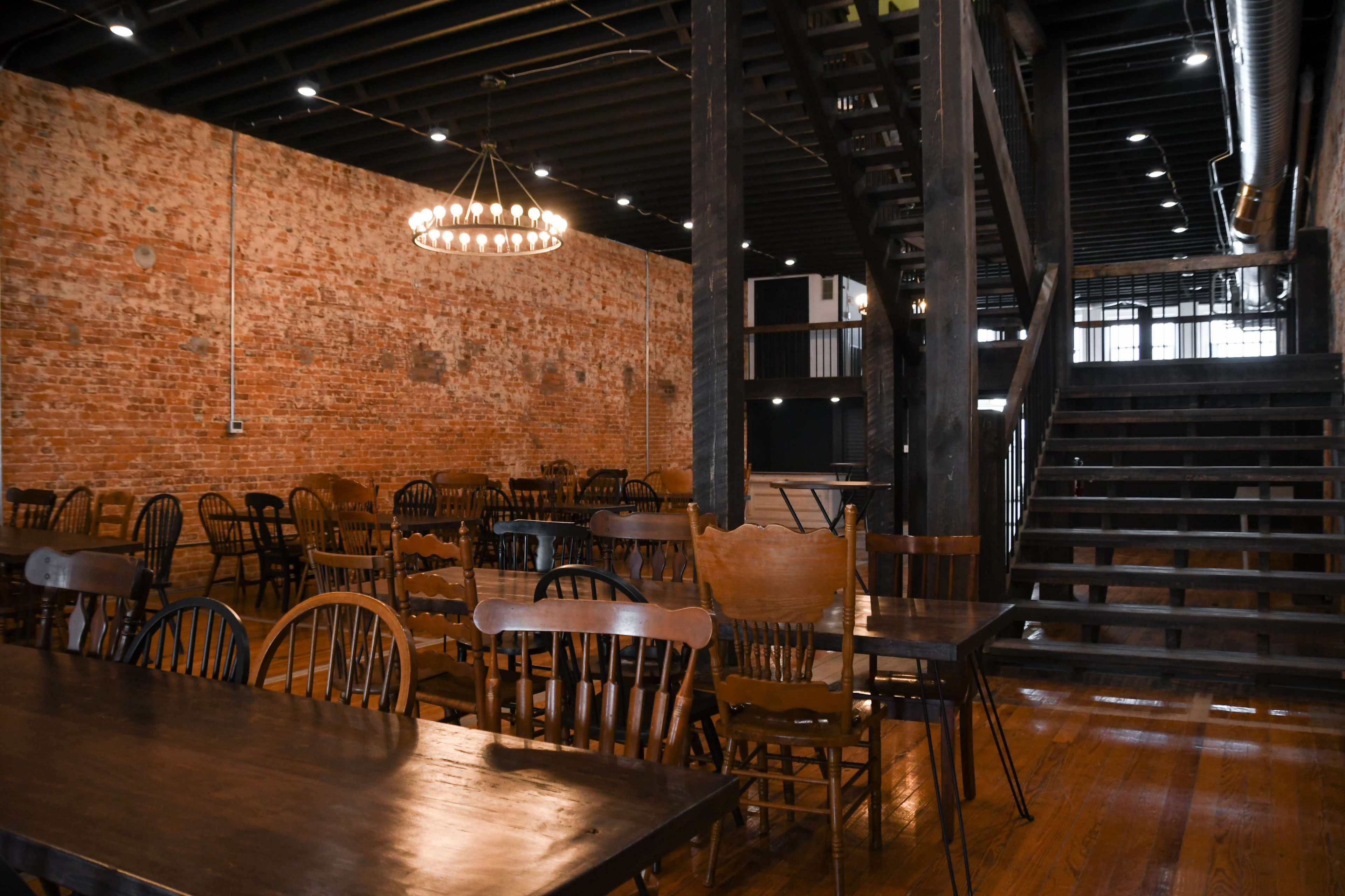 A large, empty dining area features wooden tables and chairs with exposed brick walls and a staircase leading to an upper level.