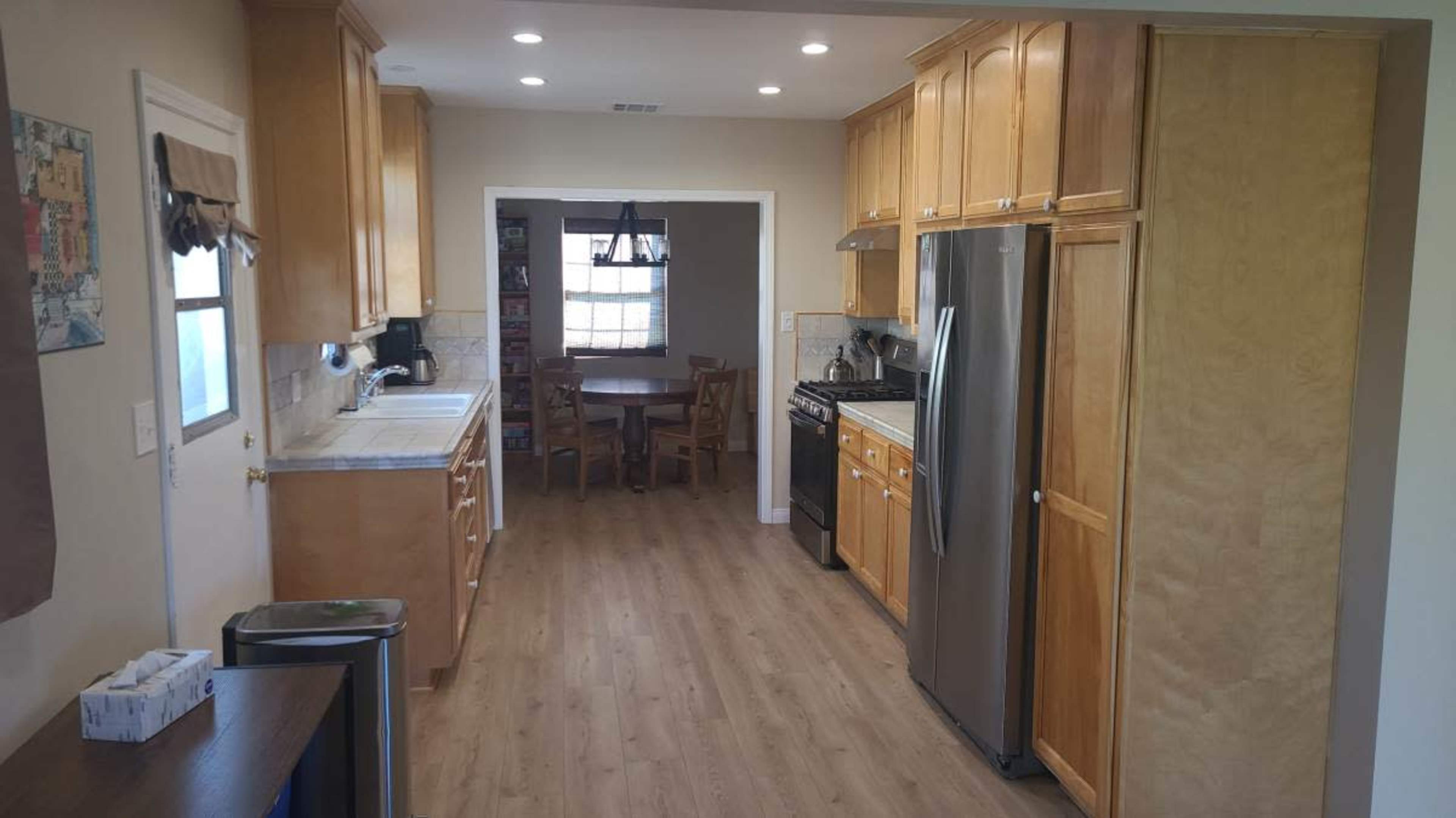 The image shows a kitchen with wooden cabinets, stainless steel appliances, and a small dining area visible in the background.