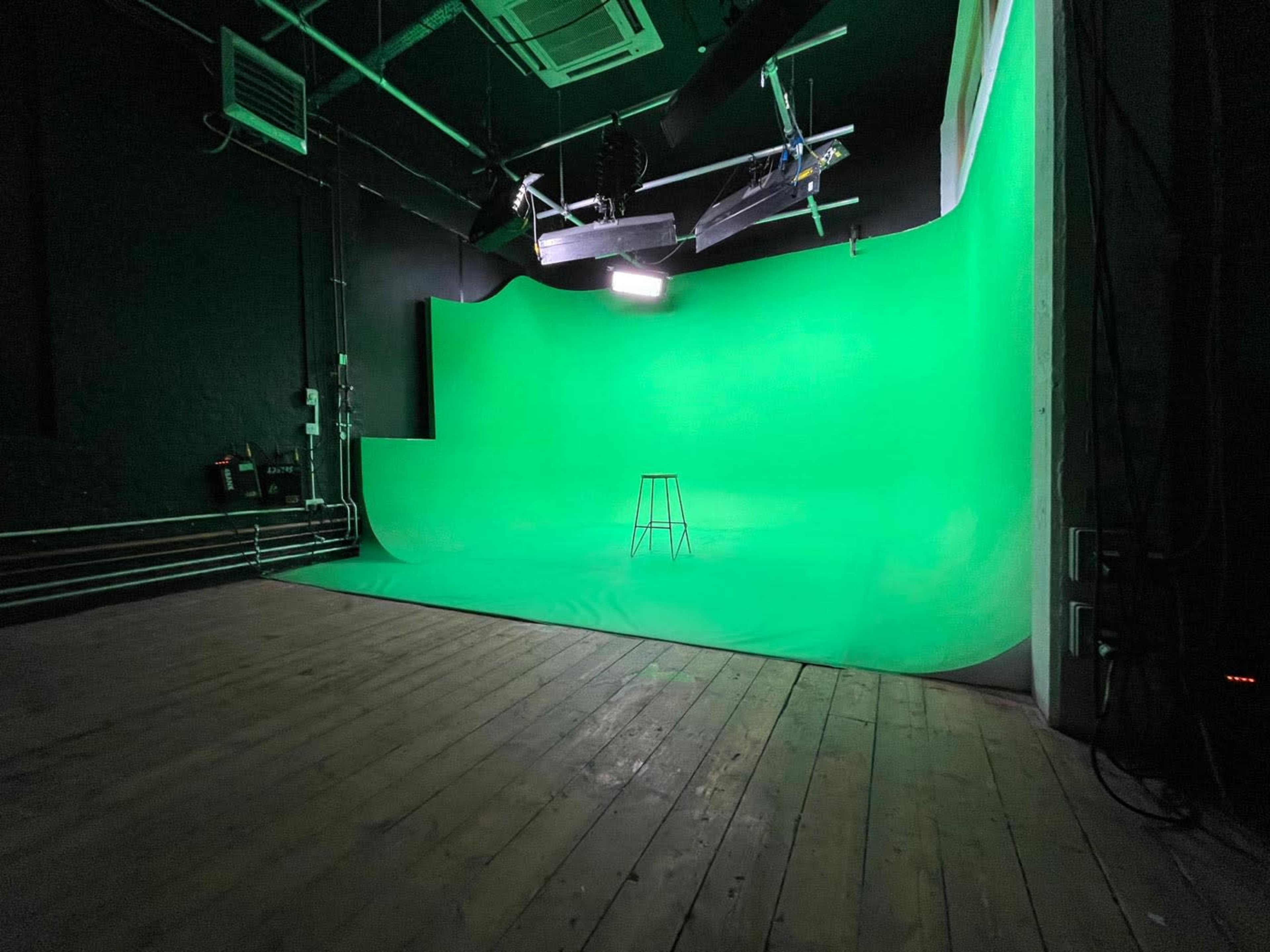 A green screen studio with a bare wooden floor and a single stool positioned in front of a large green backdrop.