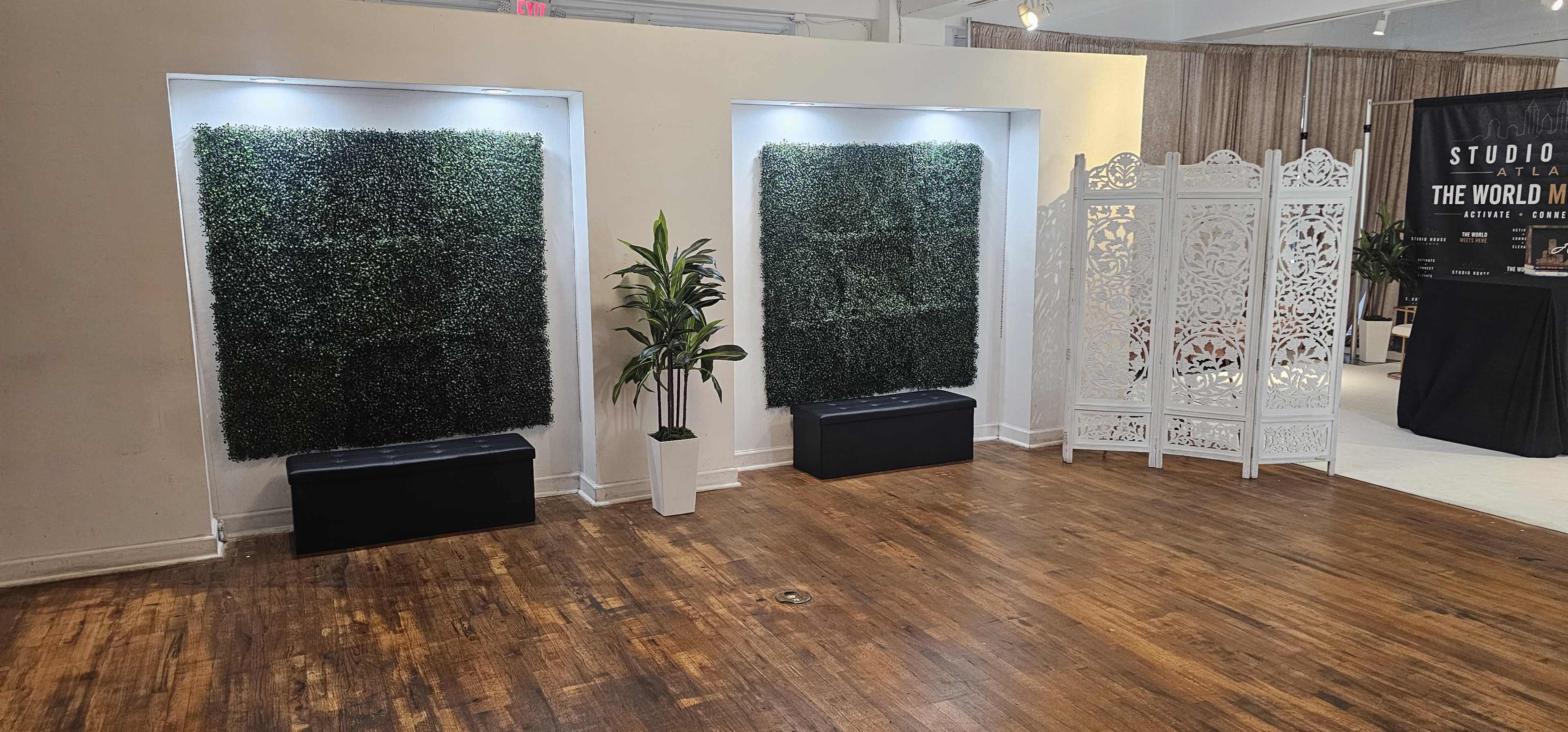 The image shows a spacious indoor area featuring two vertical green wall panels, a potted plant, and an ornate white folding screen, with a wooden floor and a backdrop displaying information about a studio.